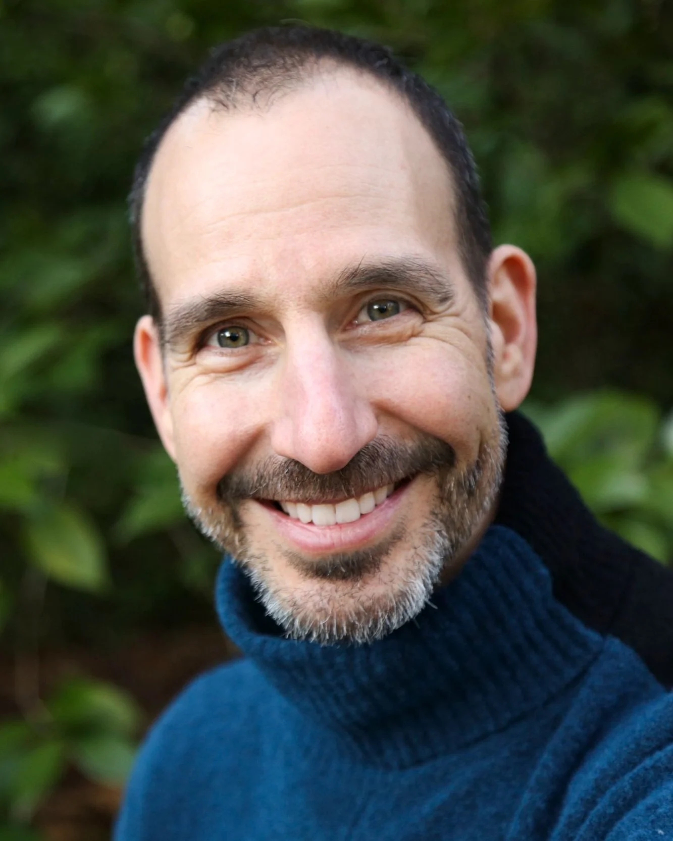 Smiling middle-aged man with a beard and short dark hair, wearing a blue turtleneck sweater, outdoors with green foliage in the background.