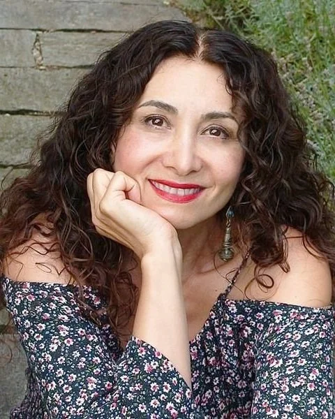 A woman with long curly dark hair smiling with her chin resting on her hand, wearing a floral off-the-shoulder top, with a stone wall and greenery in the background.