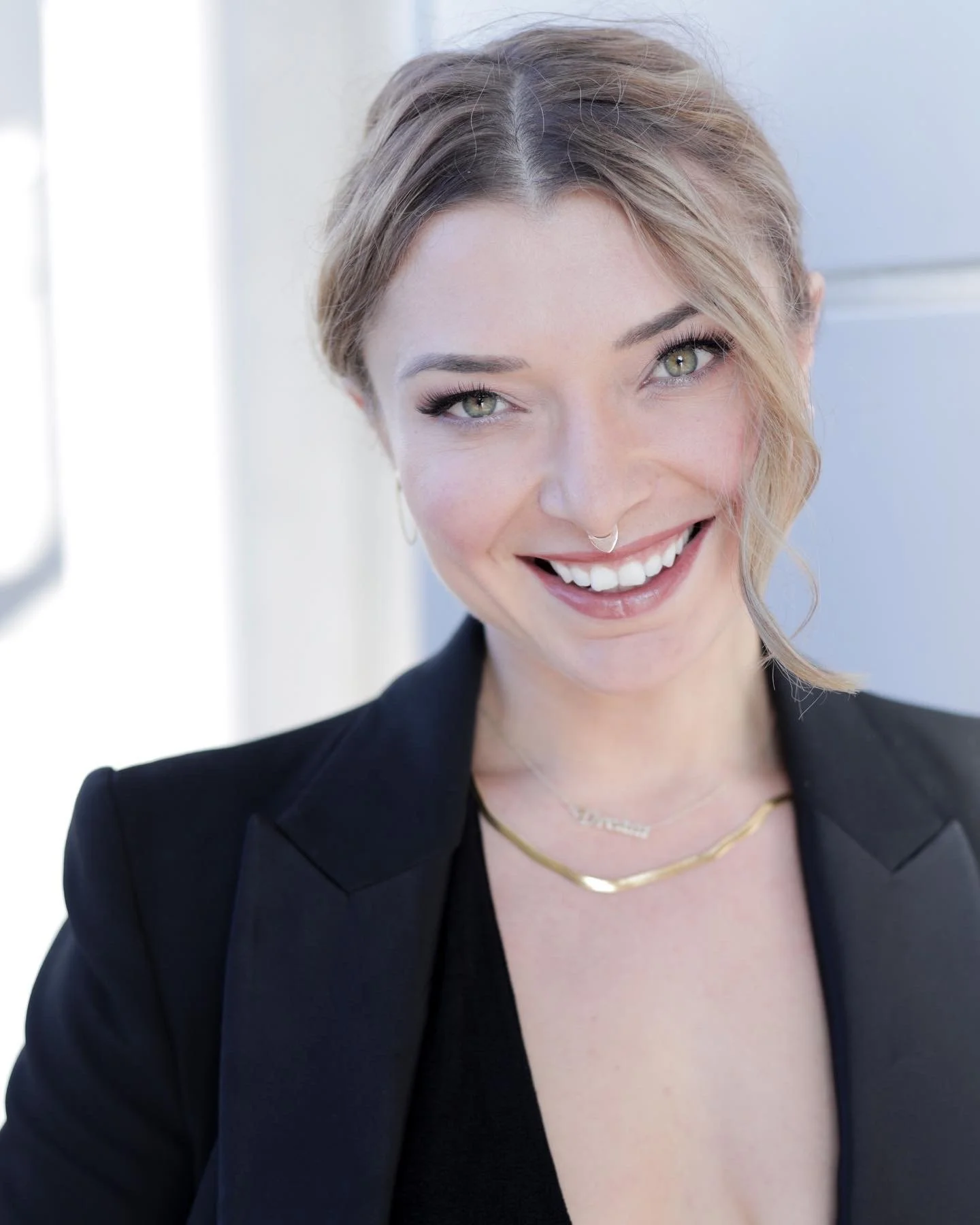 A woman with light brown hair styled in loose waves, wearing a black blazer, gold necklaces, a septum nose ring, and makeup, smiling in an indoor setting with a bright background.