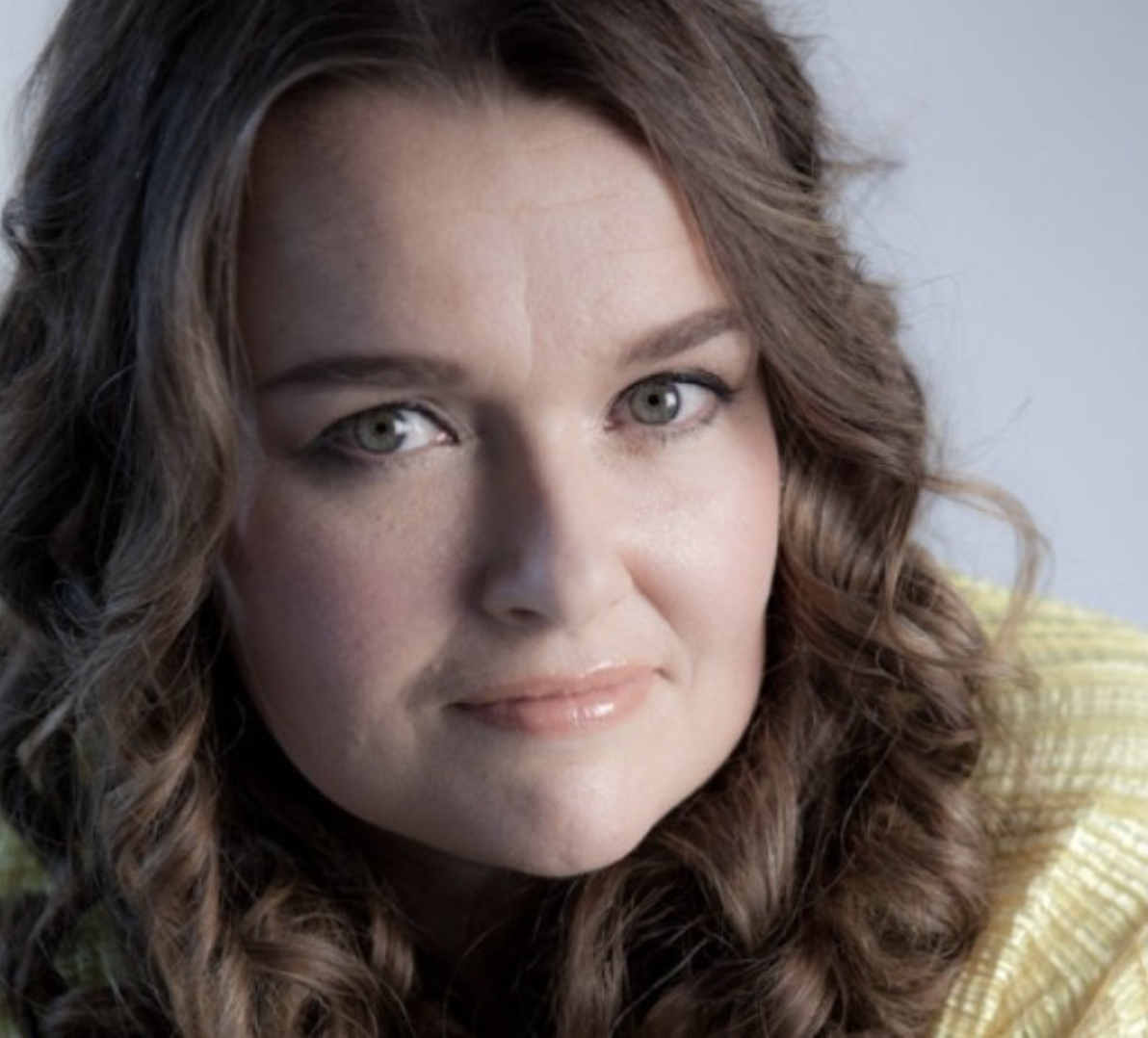 Close-up of a woman with long curly brown hair and gray eyes, wearing a yellow sweater.