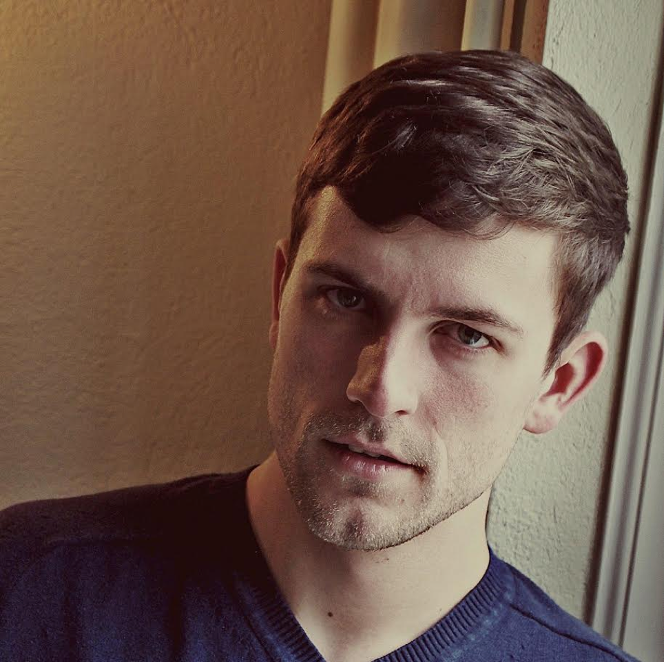 A young man with short brown hair and light stubble, wearing a navy blue shirt, looking at the camera with a neutral expression, standing next to a window.