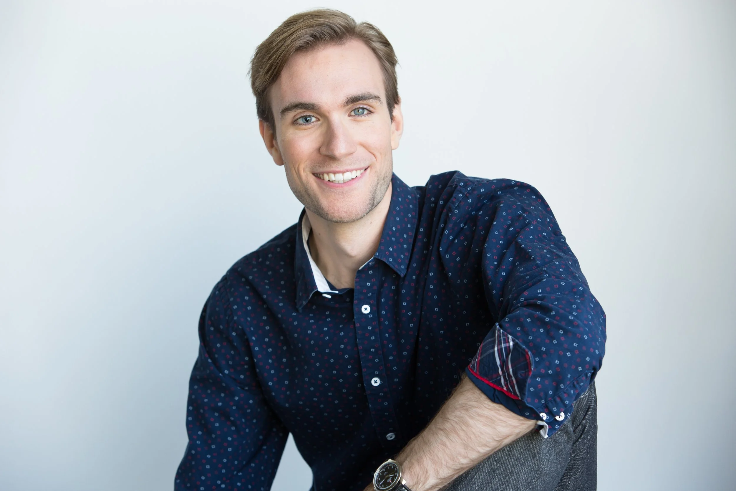 Young man with light brown hair, blue eyes, wearing a dark blue patterned shirt, smiling, against a plain white background.