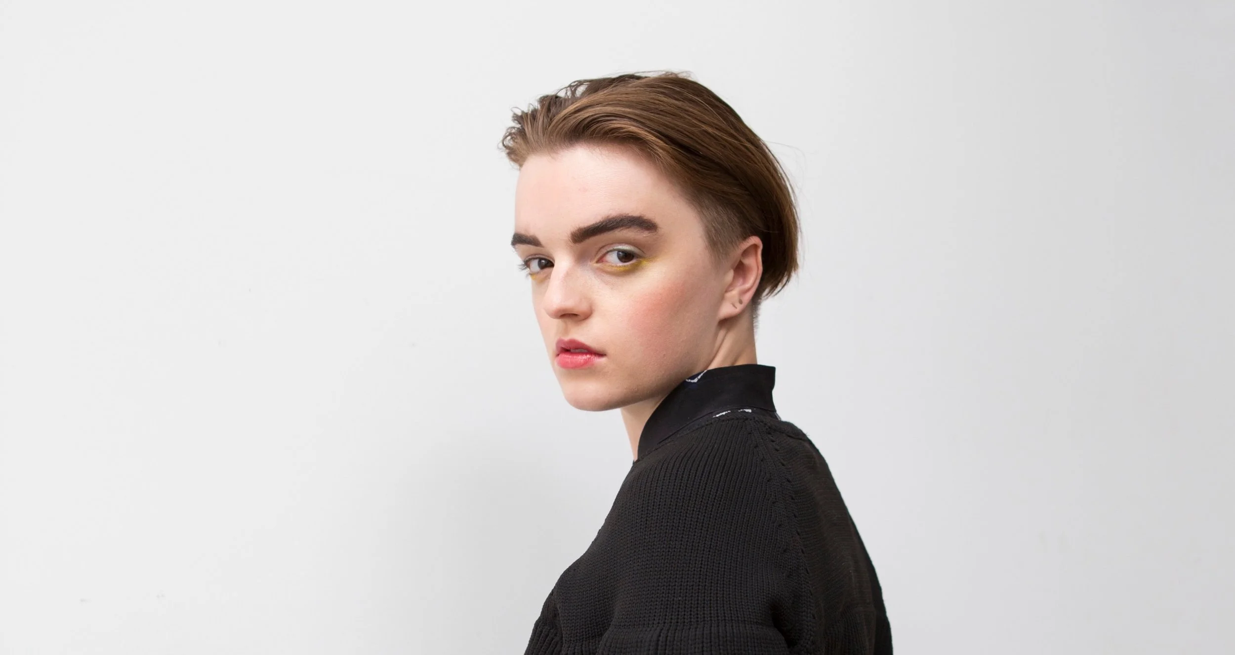 A young person with short brown hair, wearing a black top, looking over their shoulder at the camera, against a plain white background.
