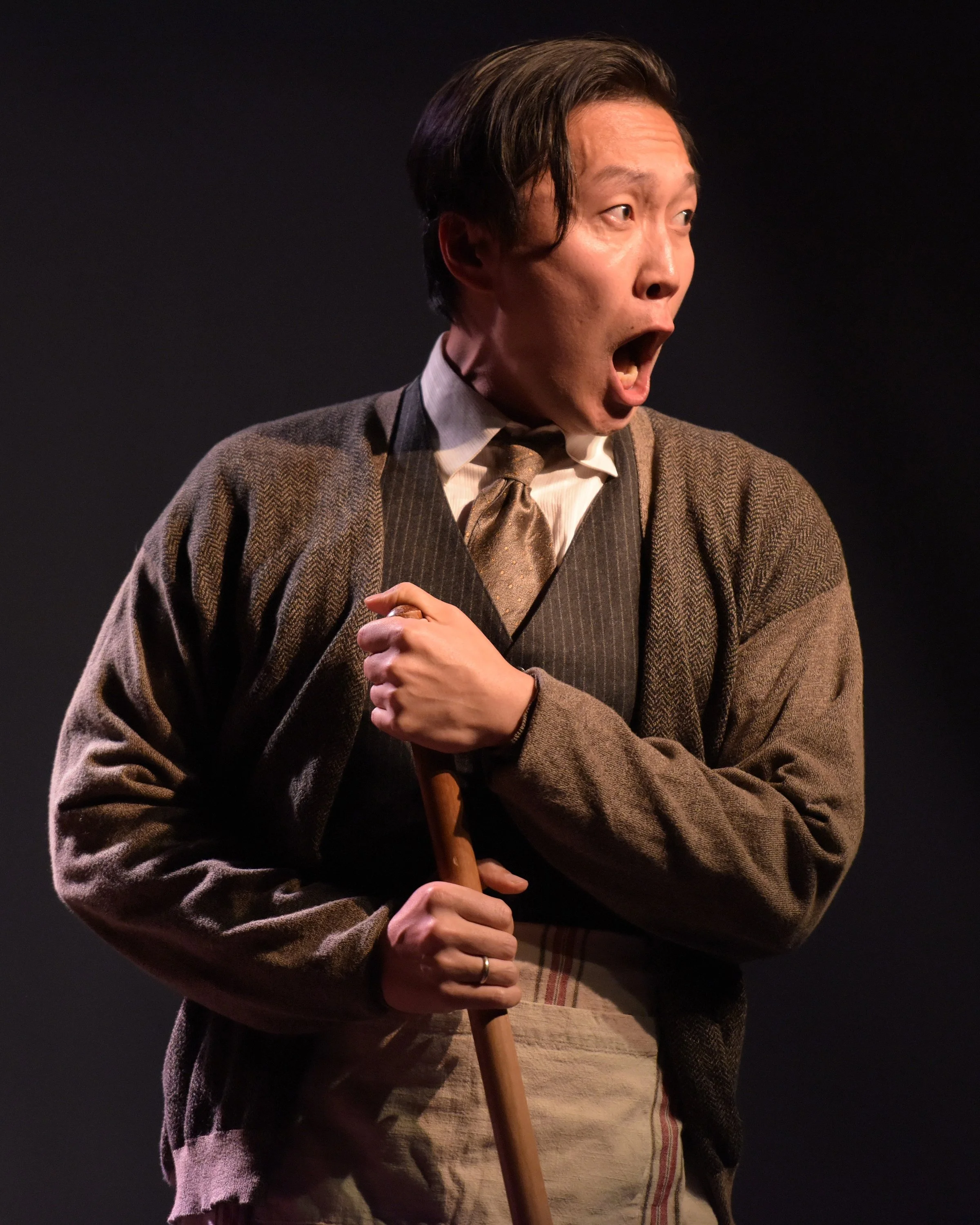 A man wearing a brown cardigan, tie, and apron, holding a broom and looking surprised or shocked.