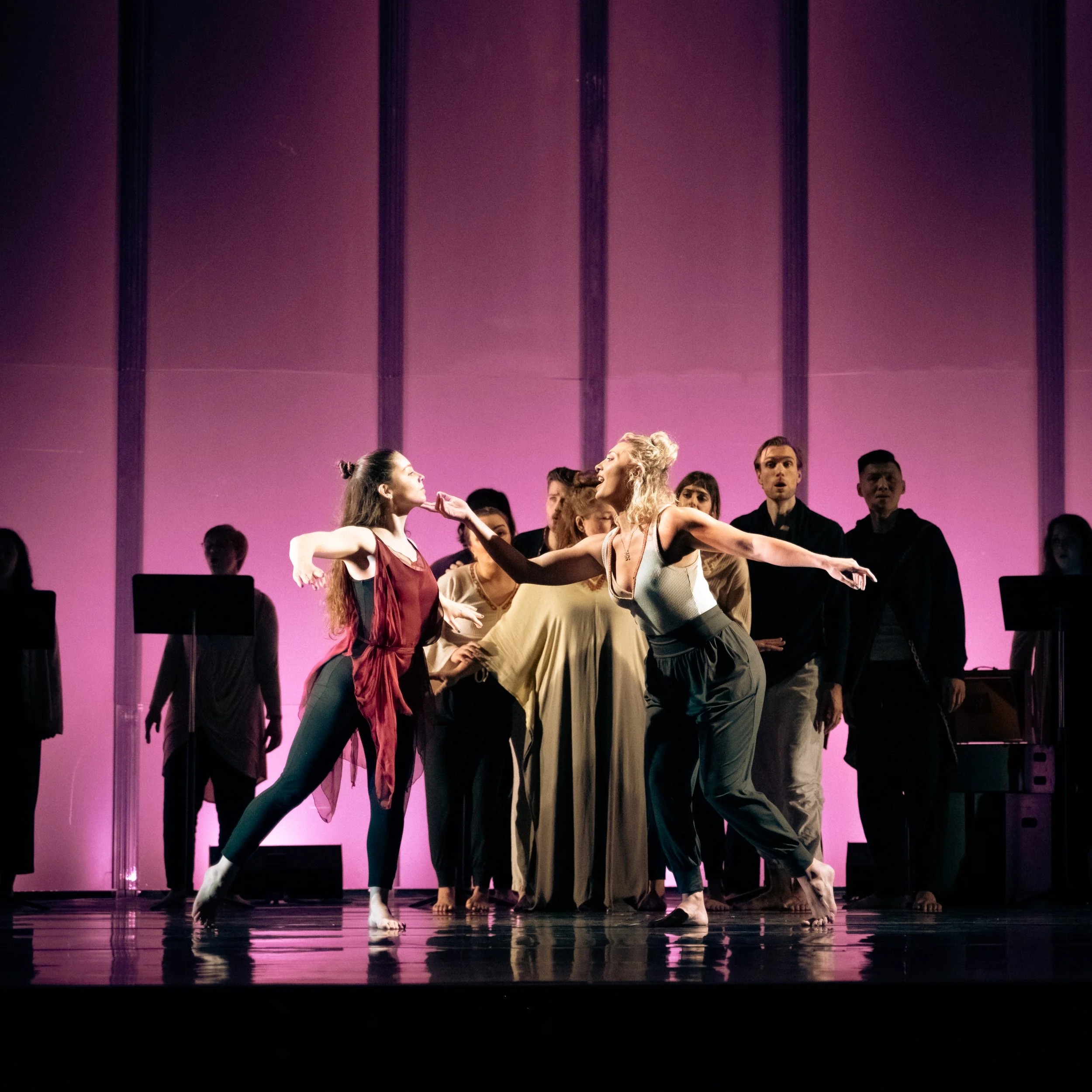 Performers on stage during a dance performance, with two women in the foreground gesturing towards each other and a group of people in the background, in front of a pink-lit backdrop.