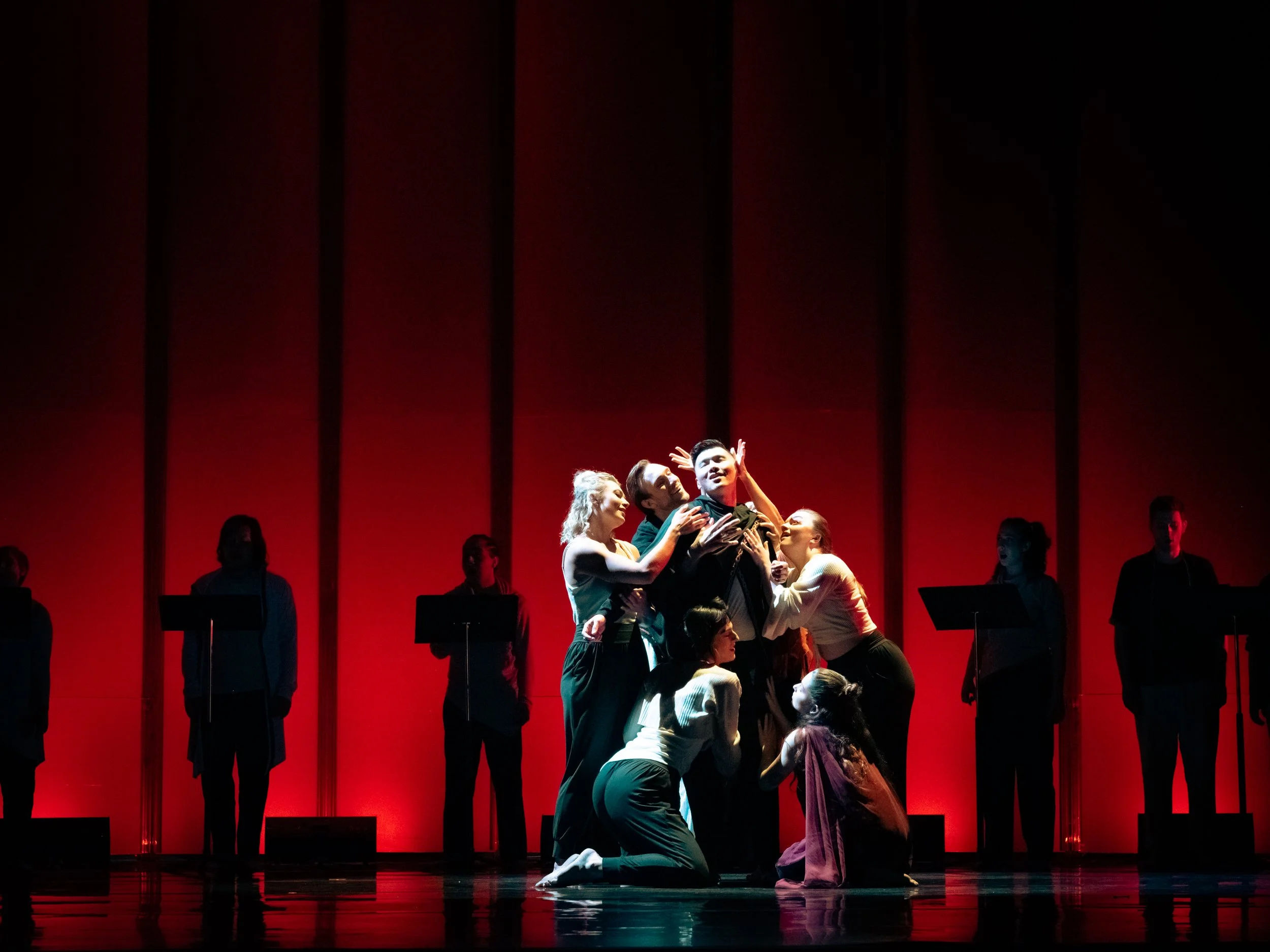 Theater scene with seven actors, six women and one man, performing on stage with a red background. The man is surrounded by women touching and engaging with him.