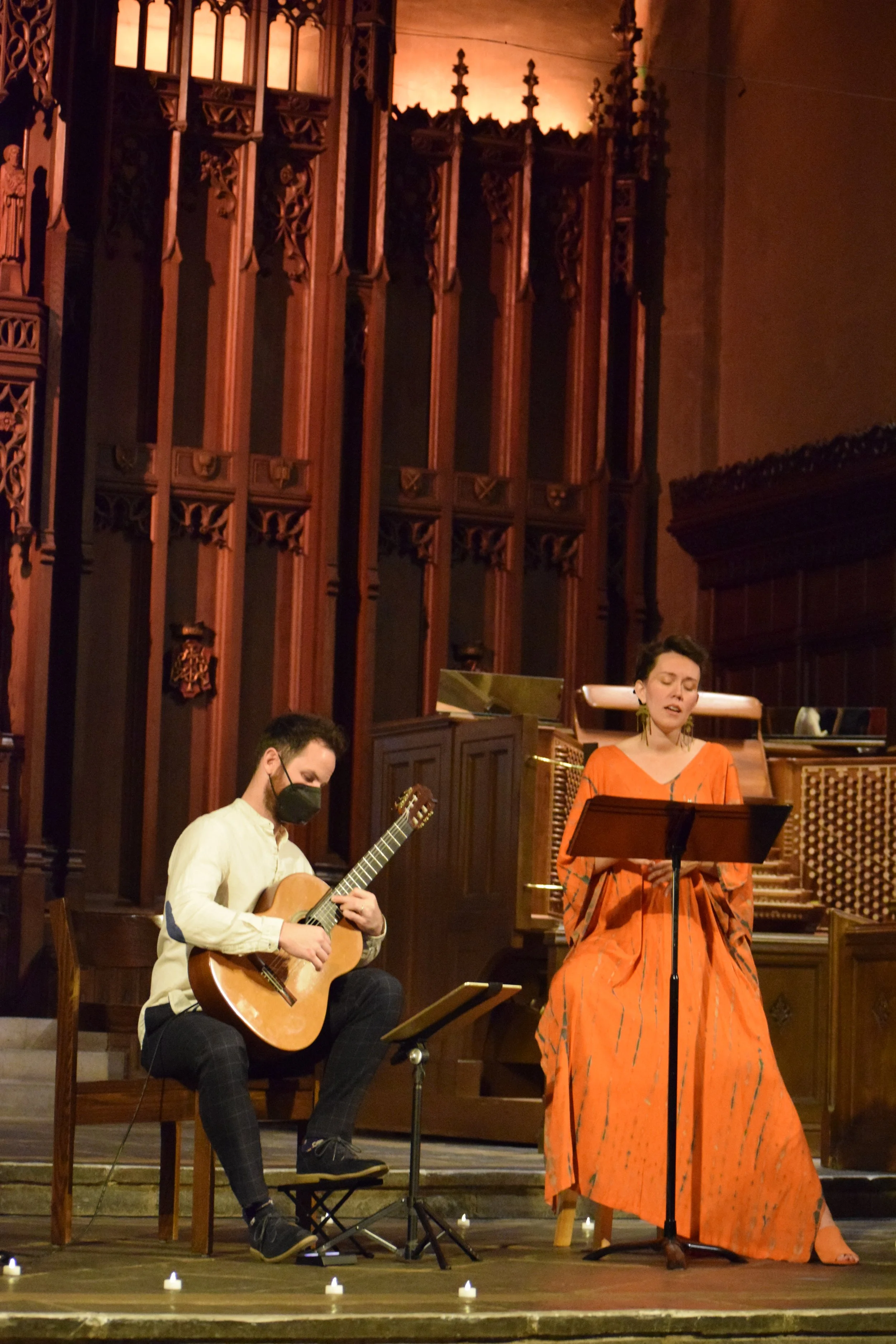 A man playing guitar and a woman singing on stage inside a church or concert hall with wood-paneled walls and large candle holders.