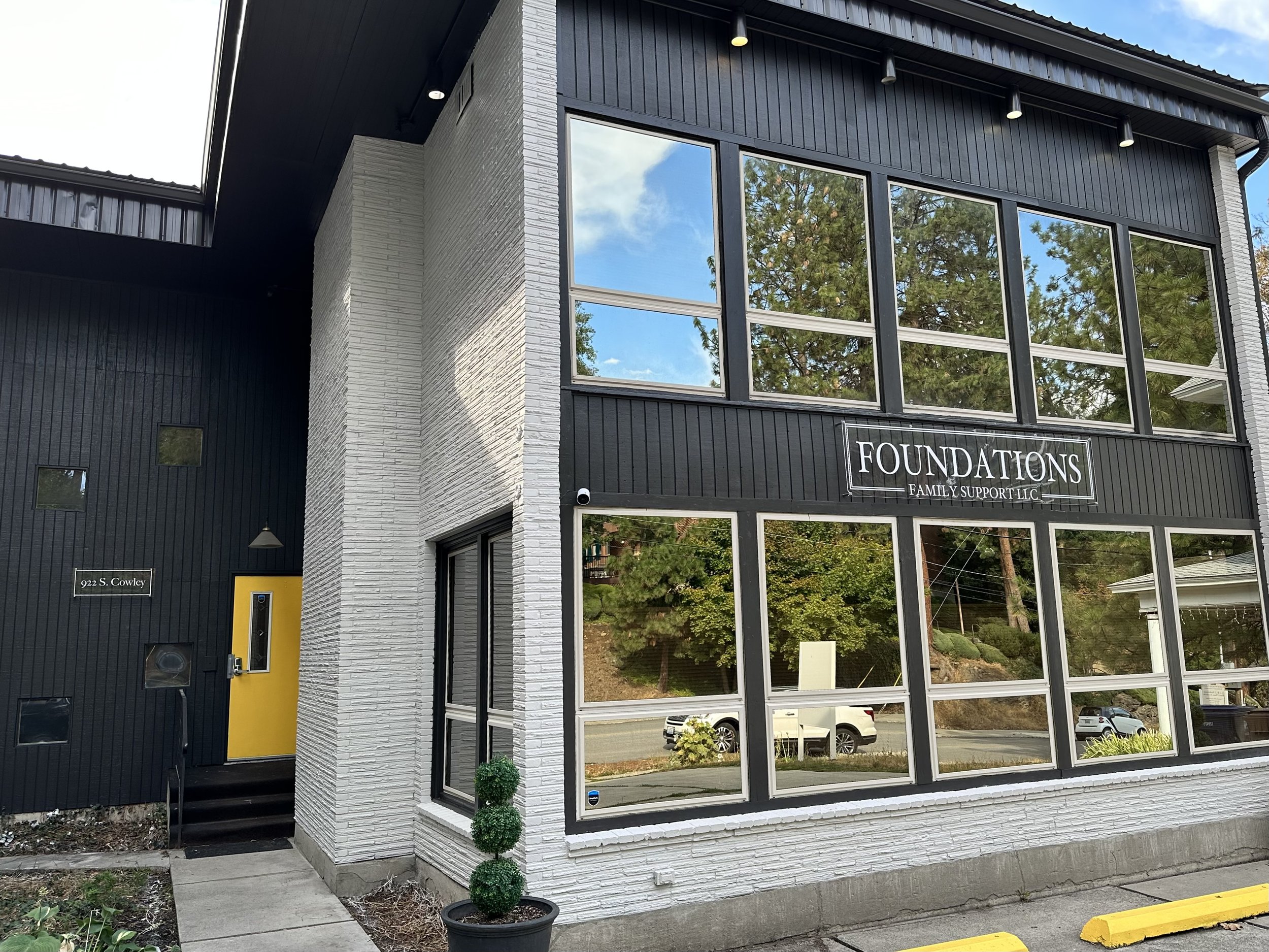 Modern building with large windows and a sign reading 'Foundations Family Support LLC.' It features a yellow door and a mix of dark and light exterior walls, located at 922 S. Cowley.