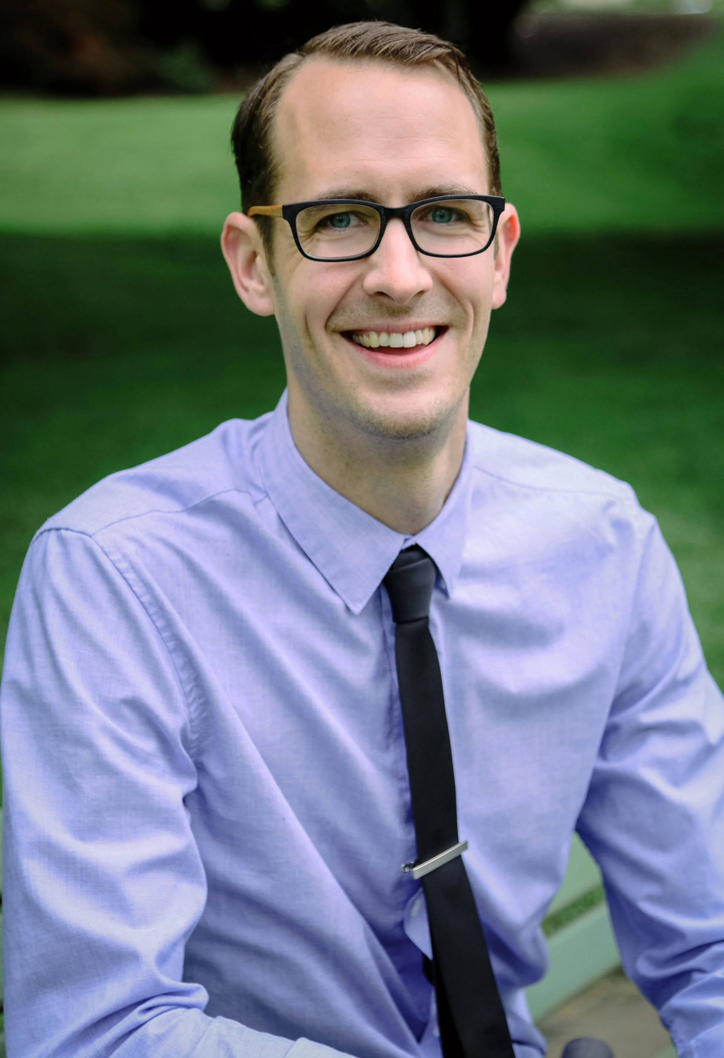 Dr. Cory wearing glasses, a blue shirt, and a black tie, smiling outdoors with a grassy background.