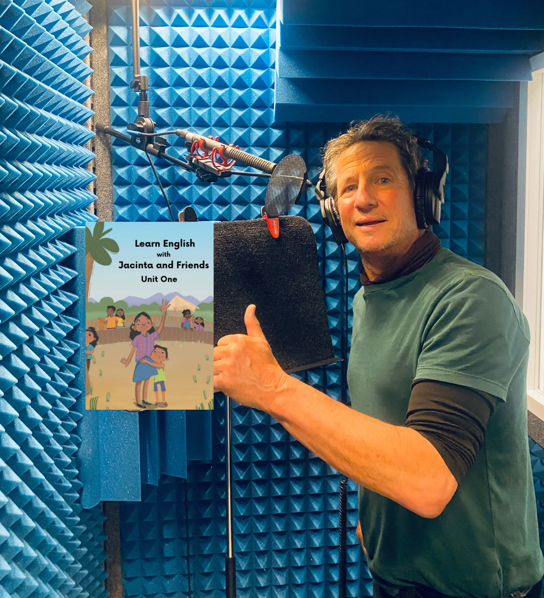 A man in a recording studio wearing headphones, standing next to a microphone with a book titled 'Learn English with Jacinta and Friends Unit One' displayed.