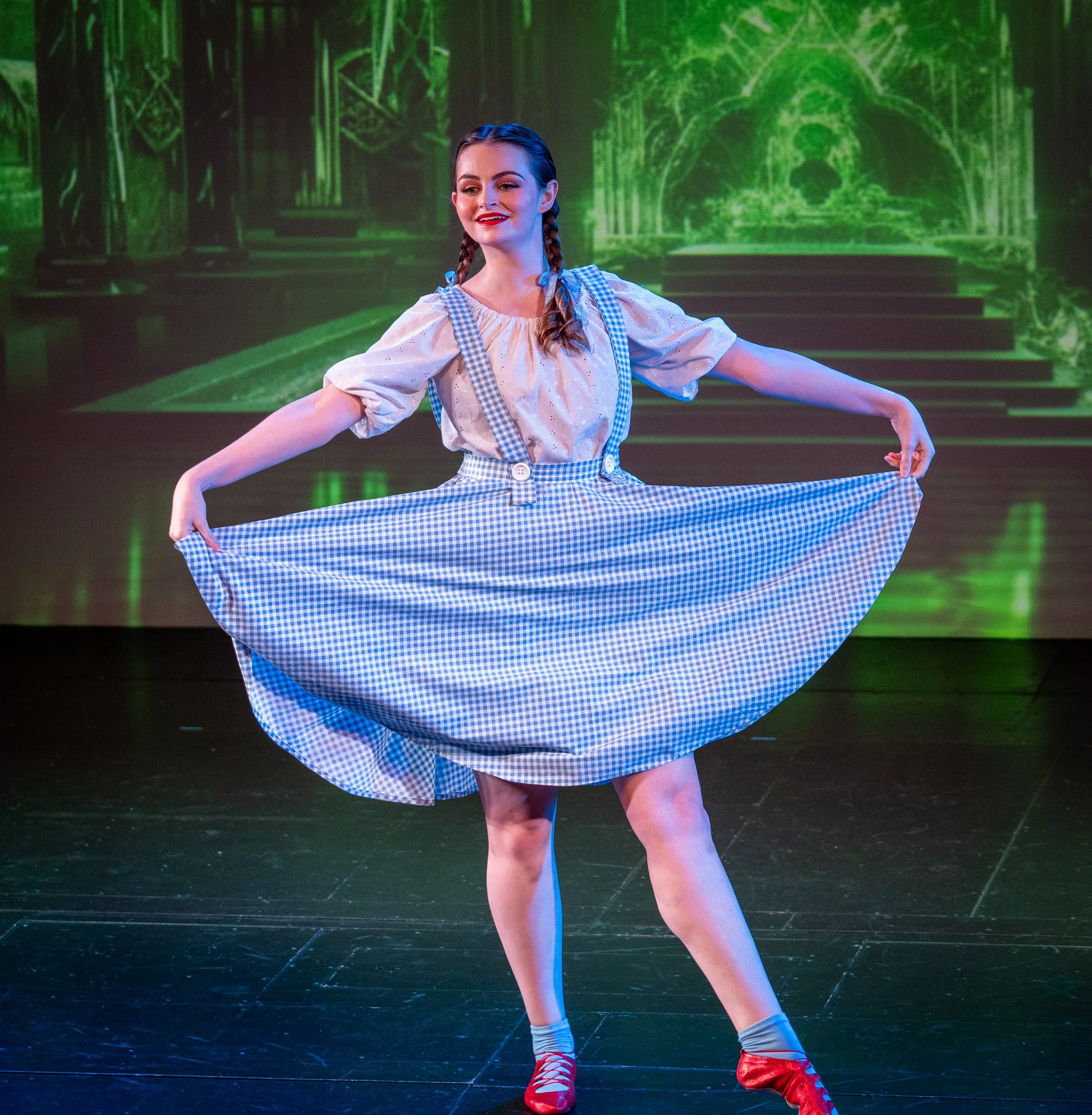 A young female dancer performing on stage with a green-lit theatrical background. She is wearing a light-colored blouse, checkered skirt, and red ballet shoes. Her hair is in two braids, and she is holding the edges of her skirt while dancing.