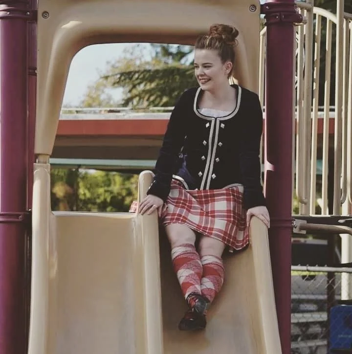 A young woman sitting at the top of a playground slide, smiling and looking to her right, outdoors with trees in the background.