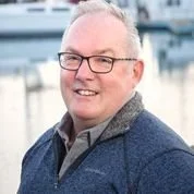A smiling middle-aged man with glasses and short gray hair, wearing a dark blue jacket, standing outdoors near a marina or waterfront.