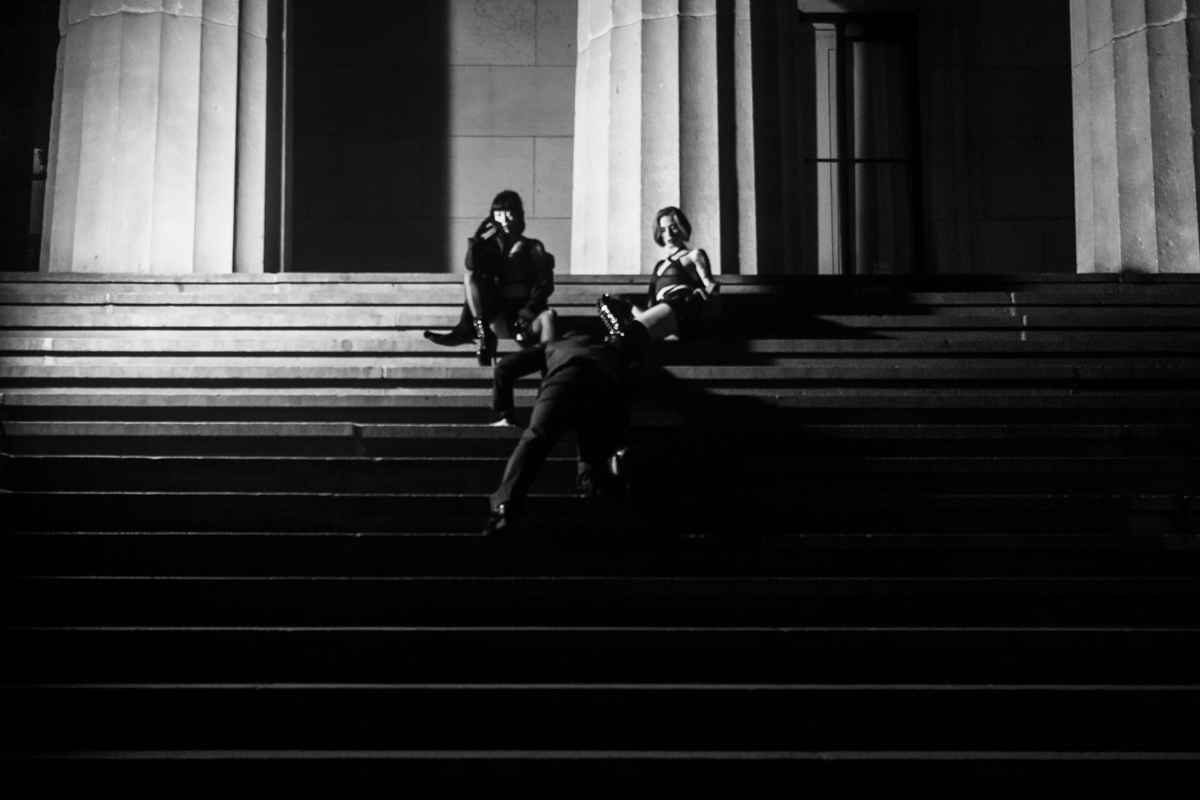 A submissive bends down to kiss the boots of Mistress Niko Flux and her wife, Mistress Sybil Fury, on a tall flight of stairs leading up to majestic columns.