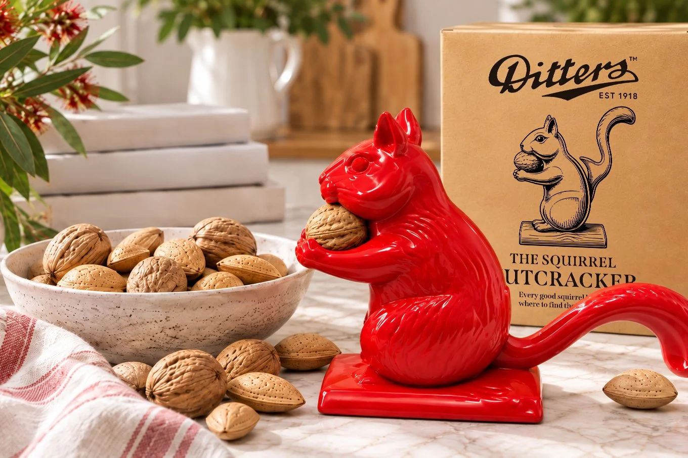 Bright lifestyle scene featuring a red Ditters Squirrel Nutcracker with its kraft gift box, surrounded by whole walnuts and almonds on a light kitchen benchtop.