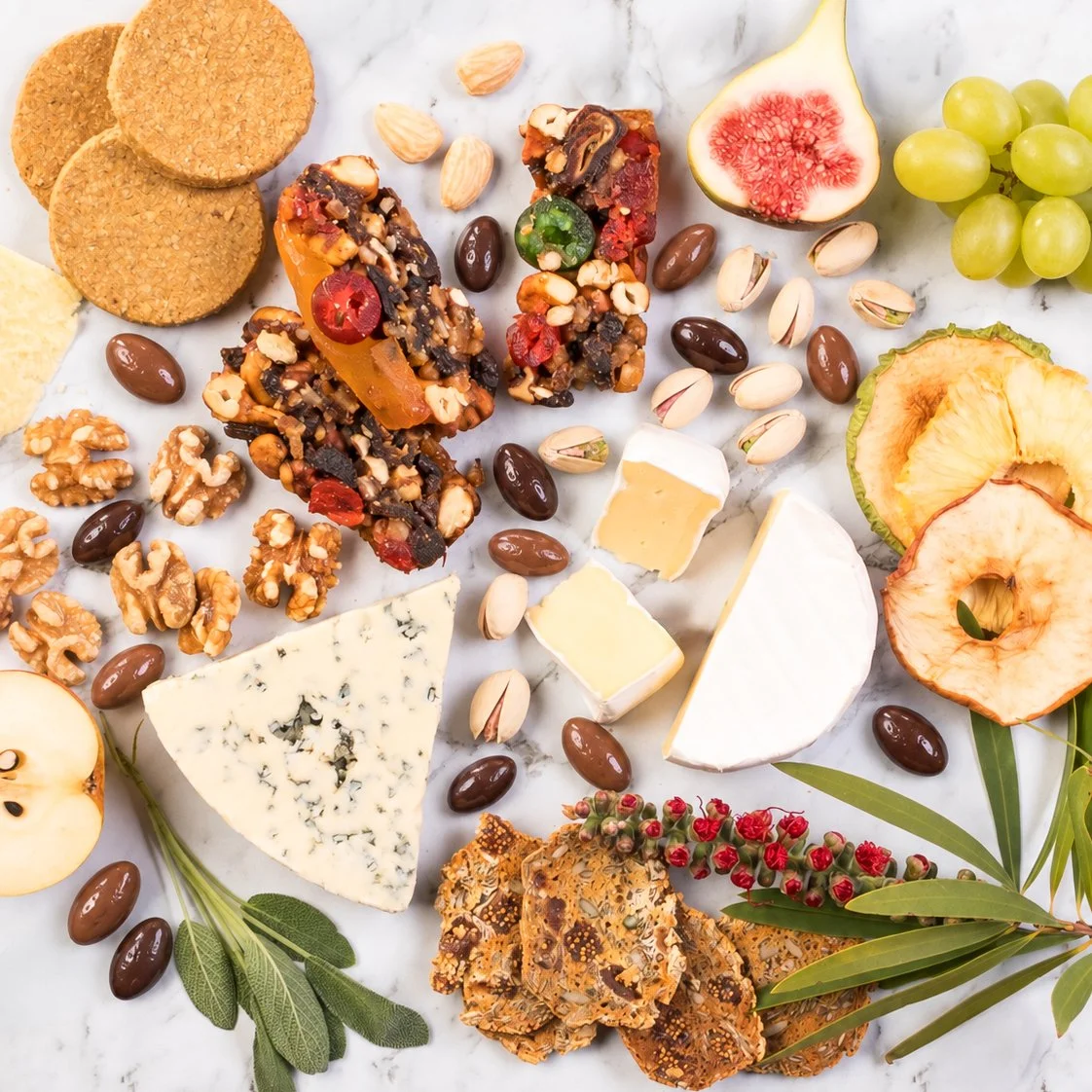 Overhead spread of cheeses, crackers, dried fruit, nuts and fresh fruit on white marble, scattered with milk and dark chocolate scorched almonds and Australian native foliage.