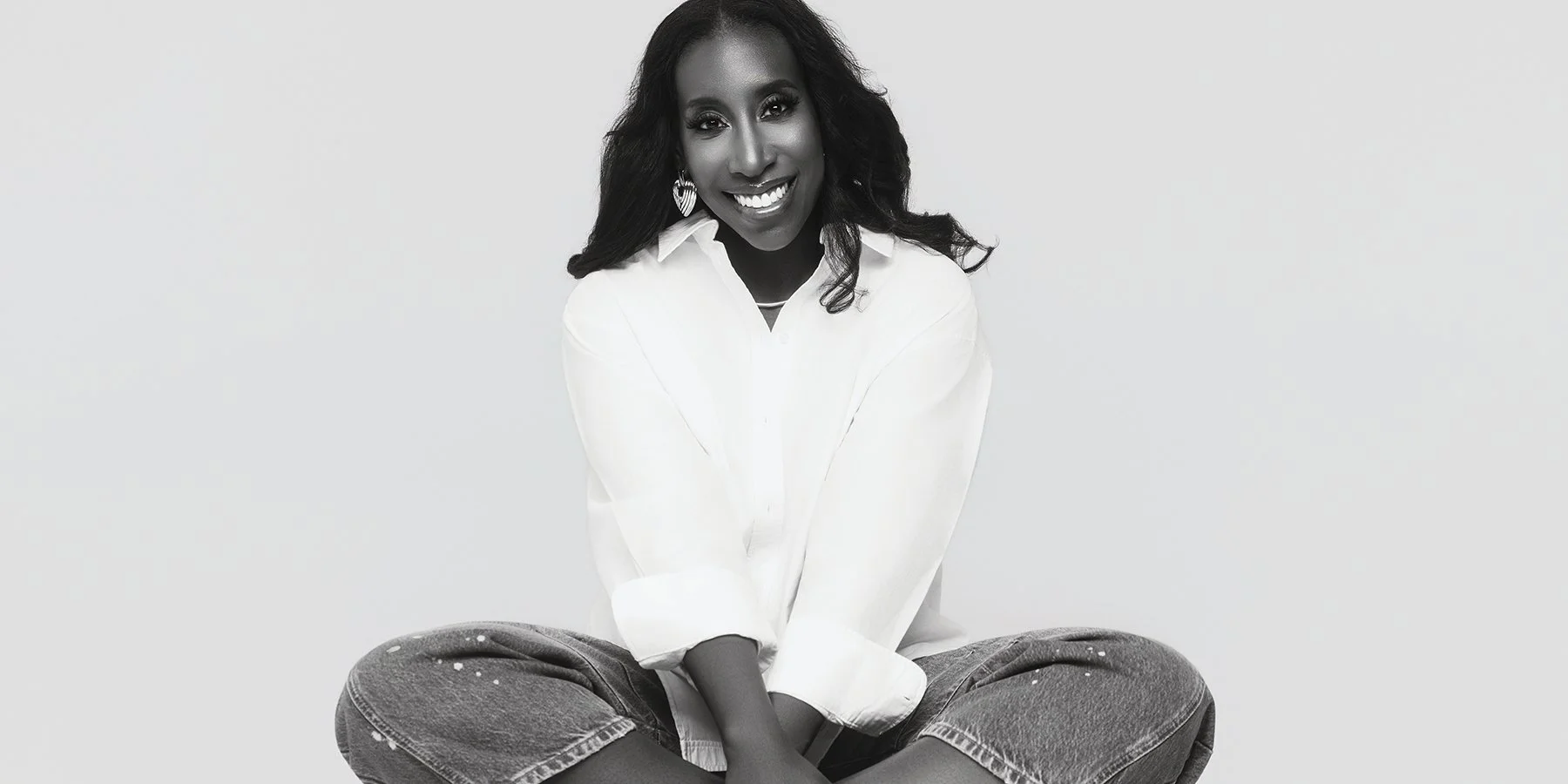 A woman with long curly hair, wearing earrings, a white shirt, and ripped jeans, sitting cross-legged and smiling at the camera.