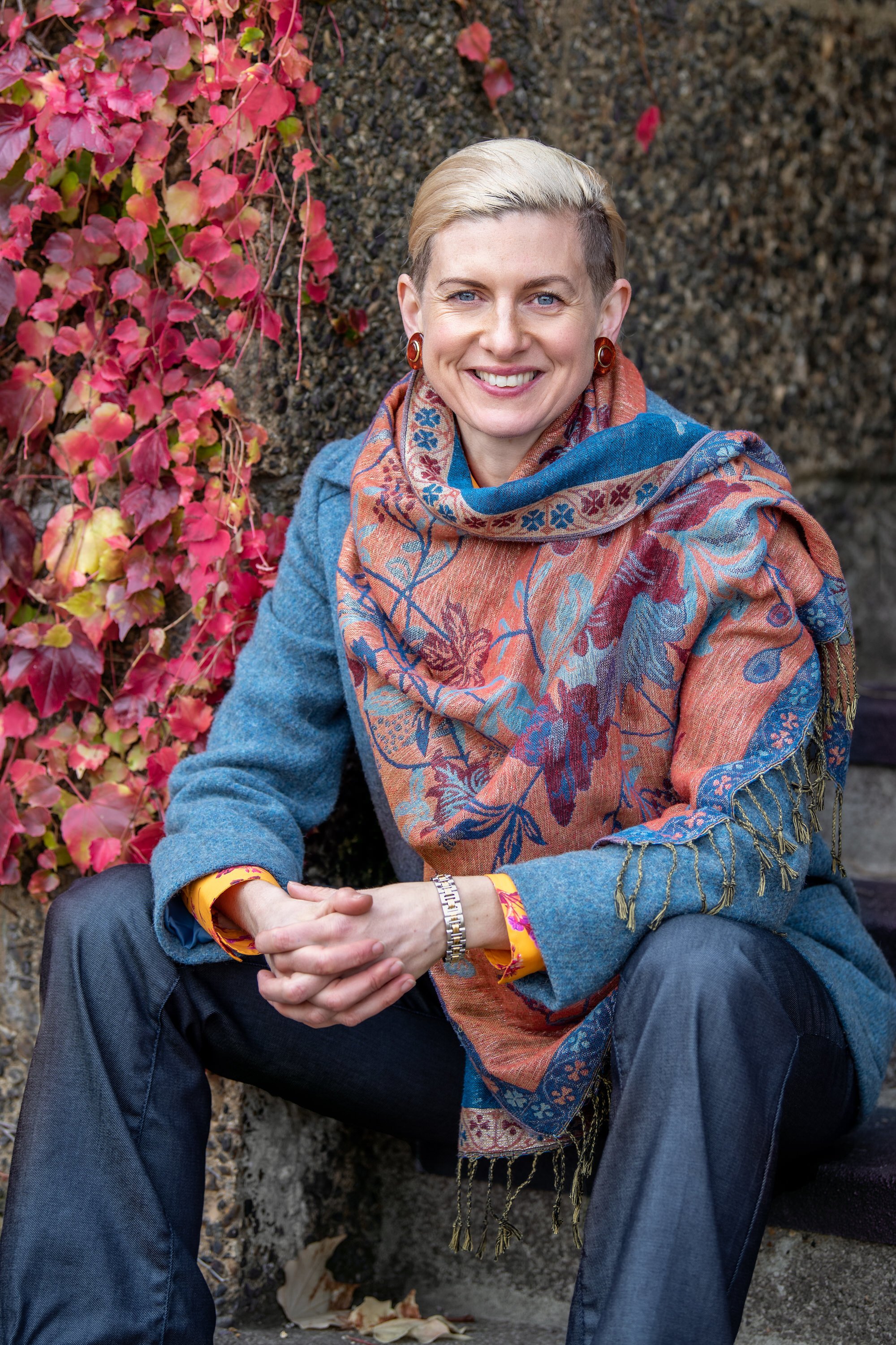 Therapist sitting on steps in winter coat with colourful scarf against an autumnal leafy backdrop. 