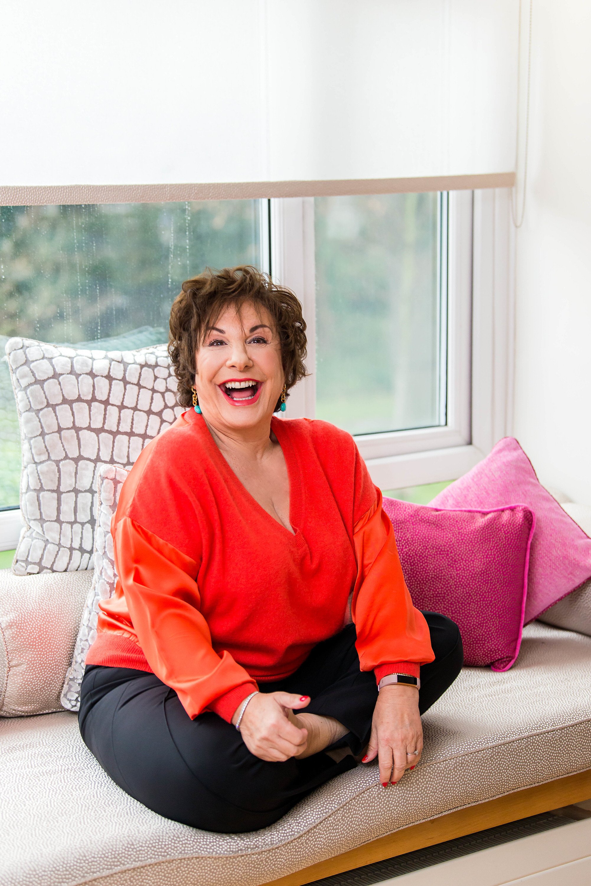  Smiling older lady in bright red jumper, sitting in a yoga pose in front of a window. 