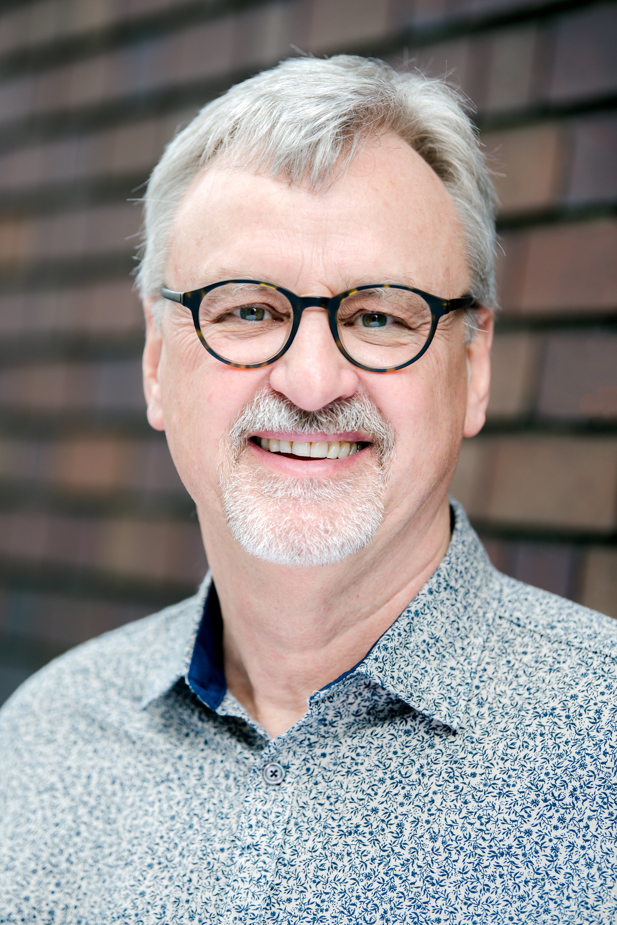  Headshot of older gentleman in glasses and shirt in front of a brick wall. 