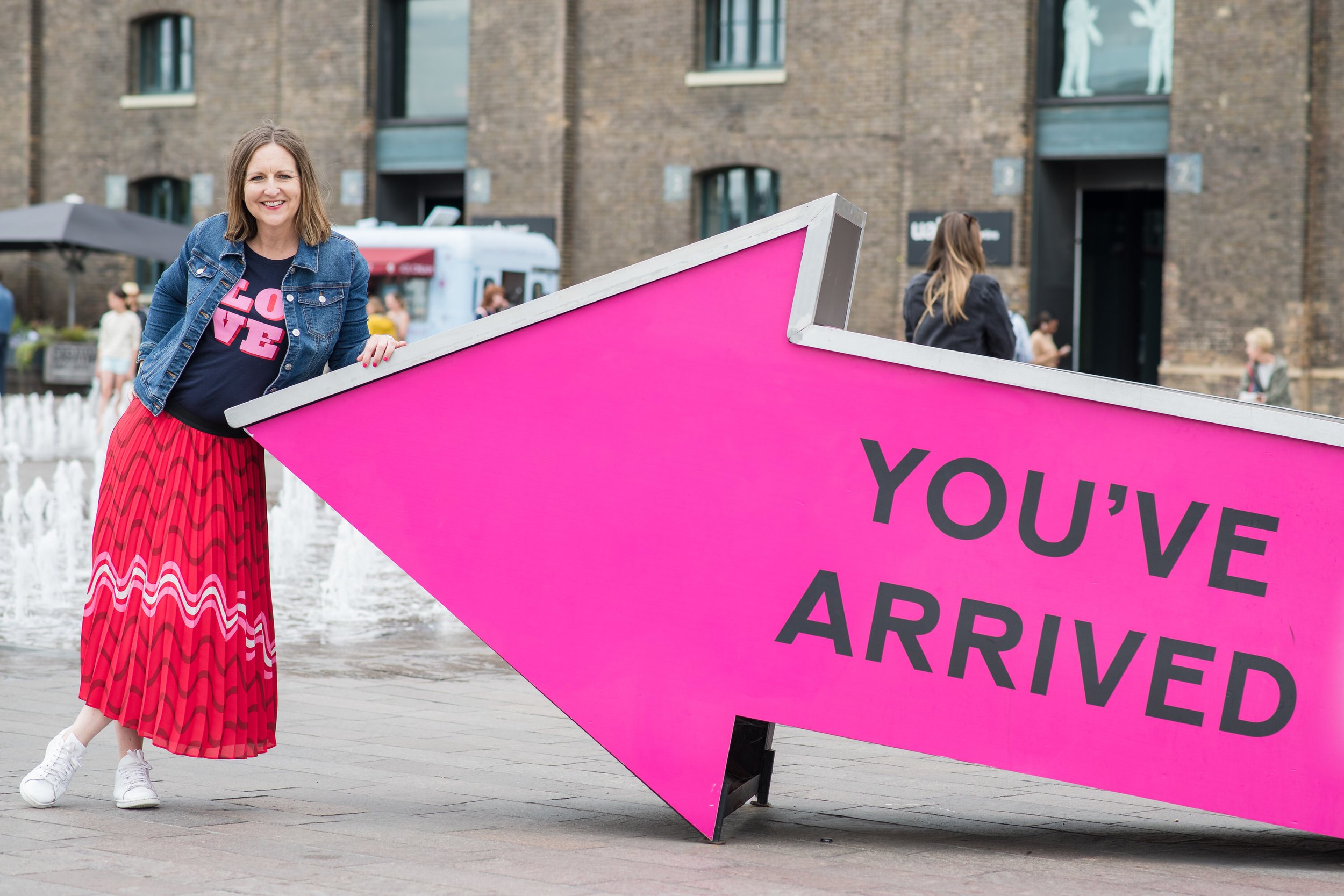  Business woman poses in front of large arrow with the words “you’ve arrived on it”  