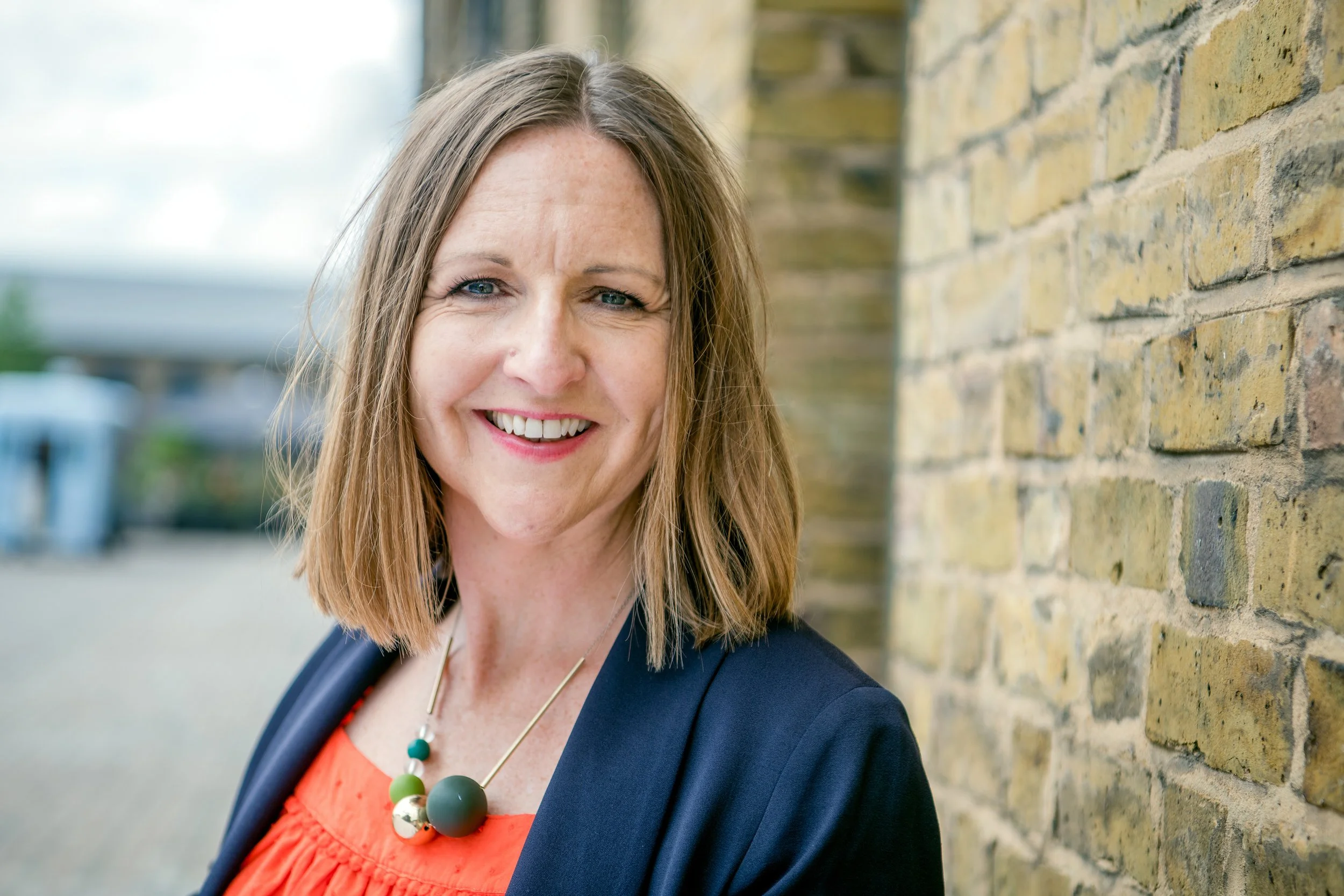 Woman in a bright orange top and blue jacket, poses for an outdoor headshot against a brick wall.