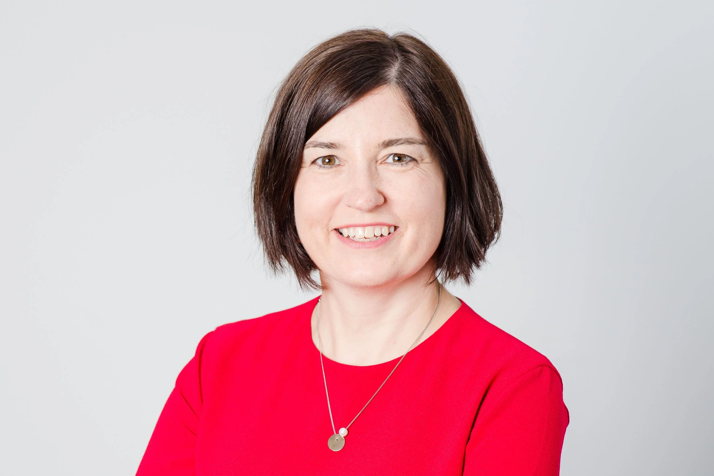 Woman in red dress smiles for a business portrait taken on a grey studio background.