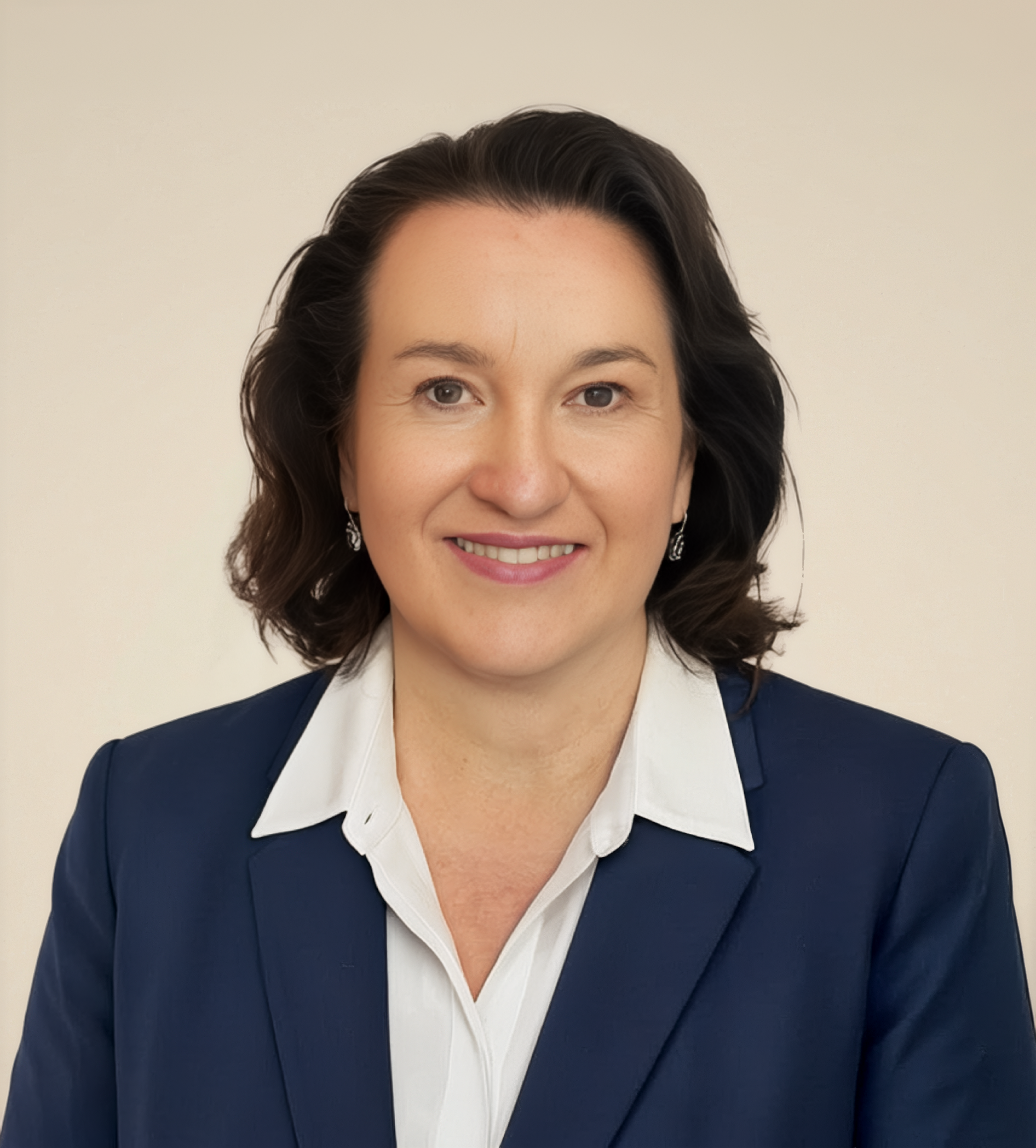 A woman with shoulder-length dark hair, wearing a dark blue blazer and white collared shirt, smiling against a plain beige background.