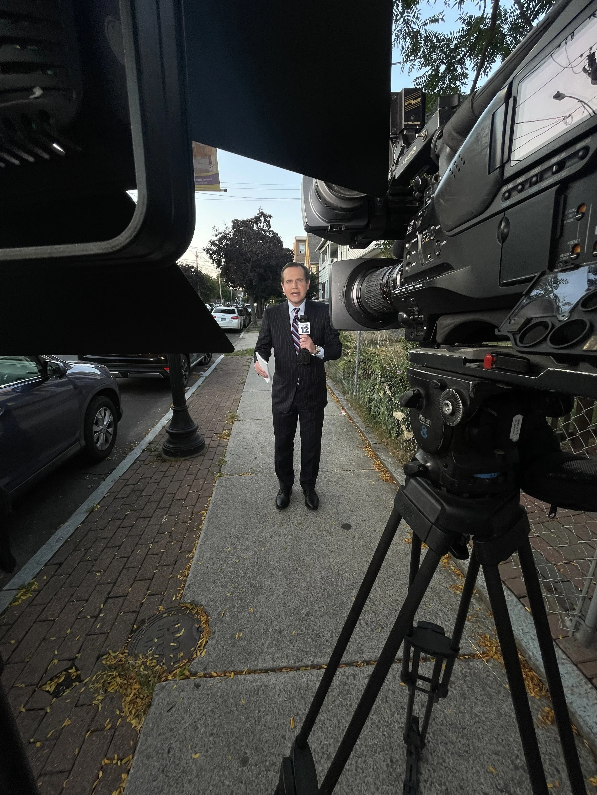 A news reporter in a suit holding a microphone with the logo 'News 12' standing on a sidewalk filming a live report. Large camera in the foreground capturing the scene. Residential street with parked cars and trees in the background.