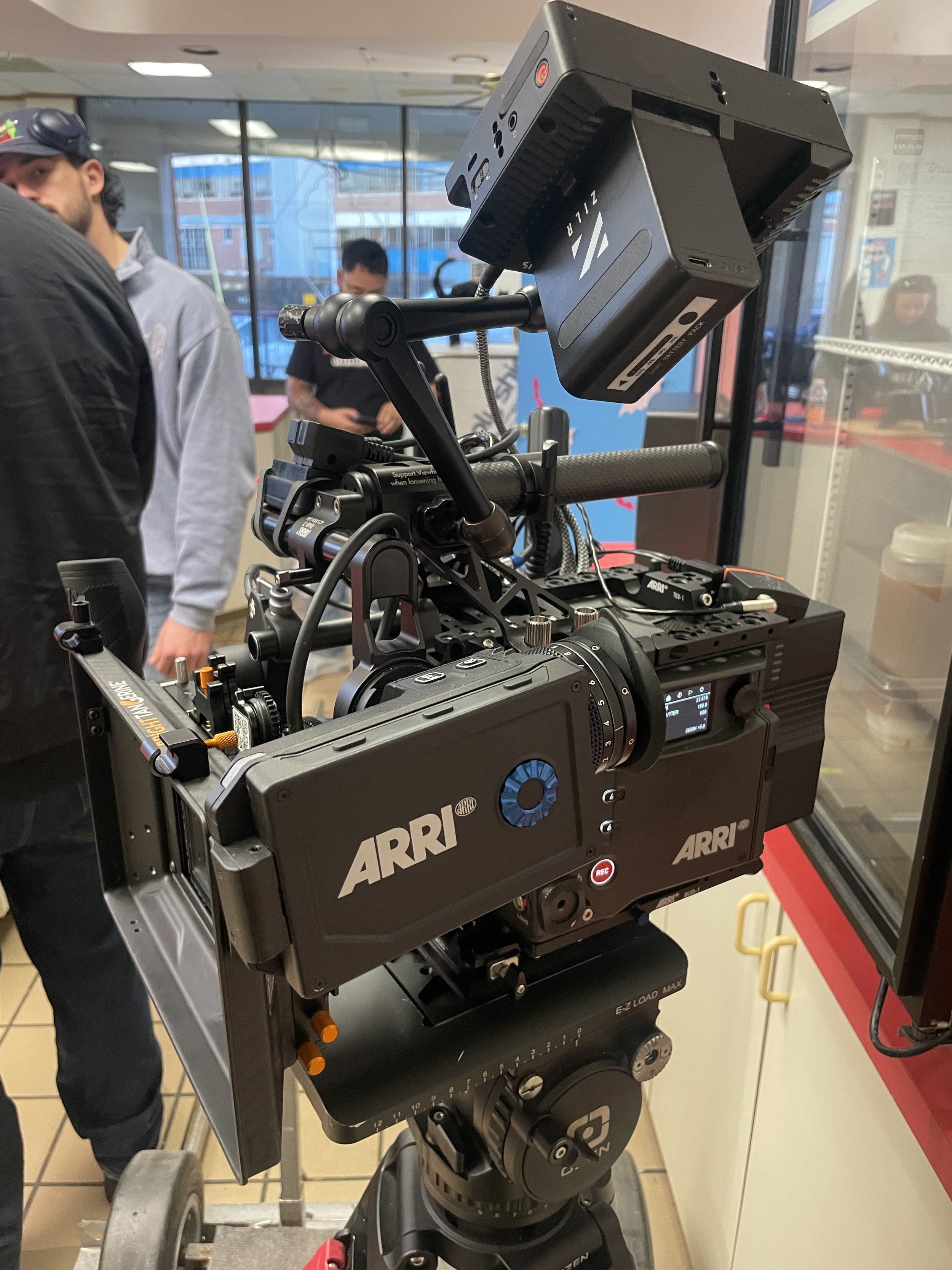 Close-up of professional video camera on a dolly, with a support monitor attached, inside a building with people in the background.