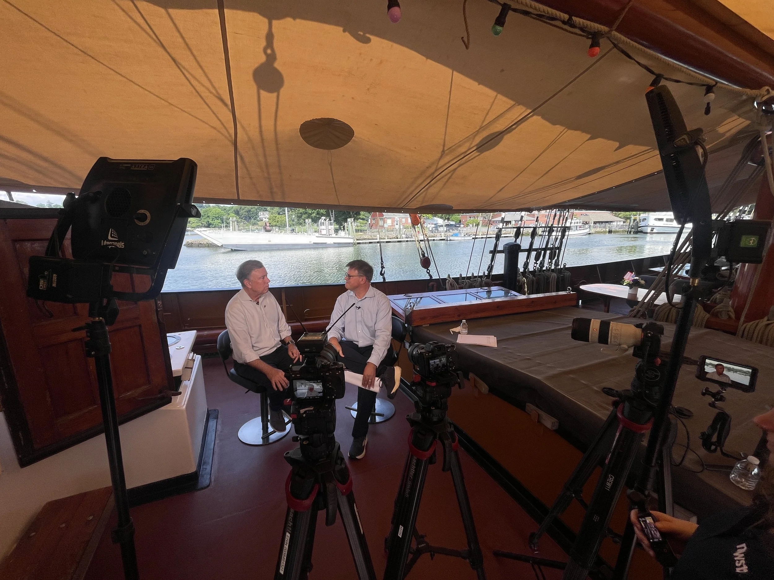 Two men are sitting and talking inside a boat with large windows, a marina and boats in the background. Cameras and equipment are set up around them, indicating an interview or recording session.