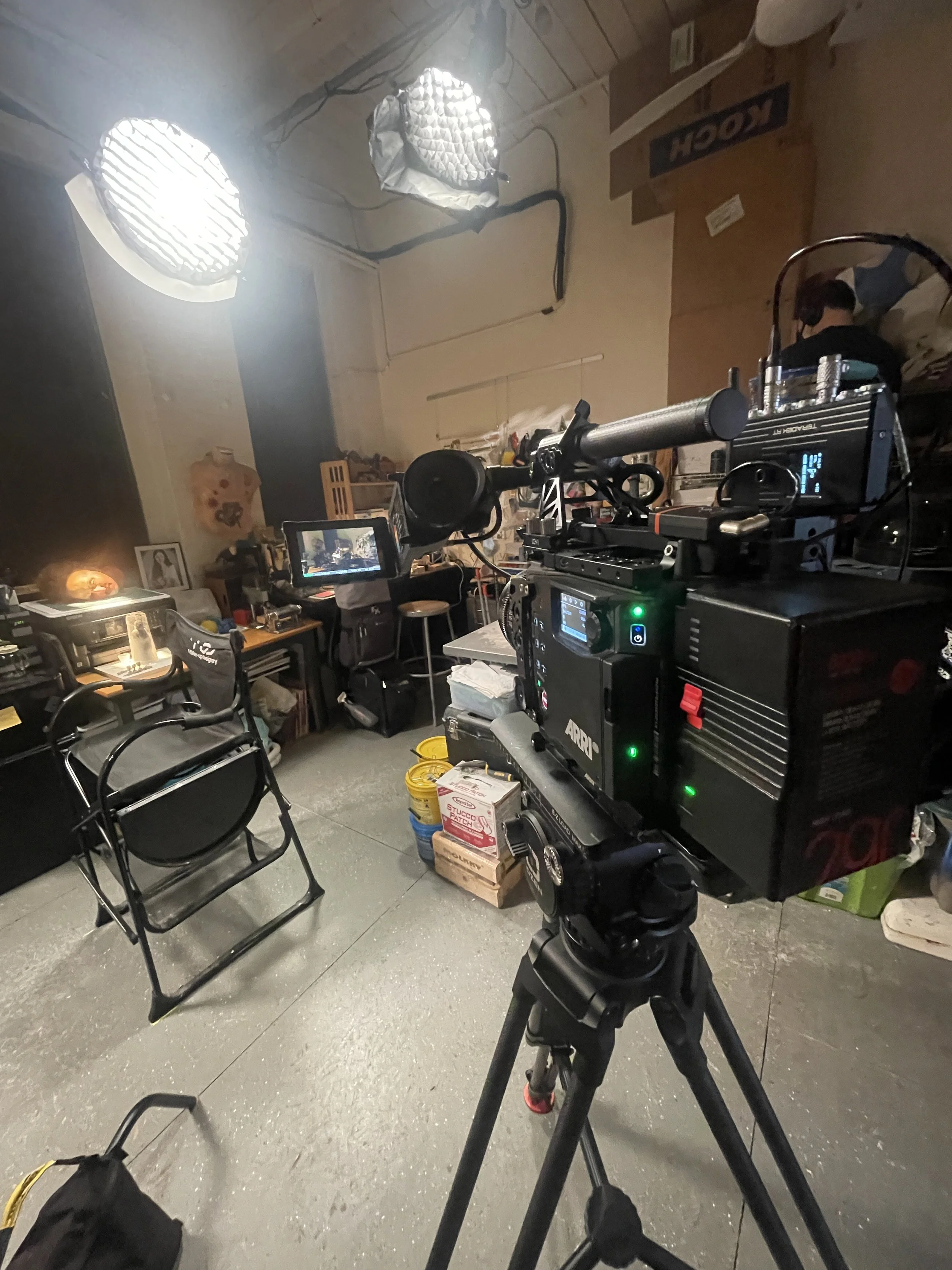 A camera on a tripod facing a cluttered room with various items including a computer, chairs, and boxes. The room has a wooden ceiling with two large studio lights and a person wearing headphones working at a desk.