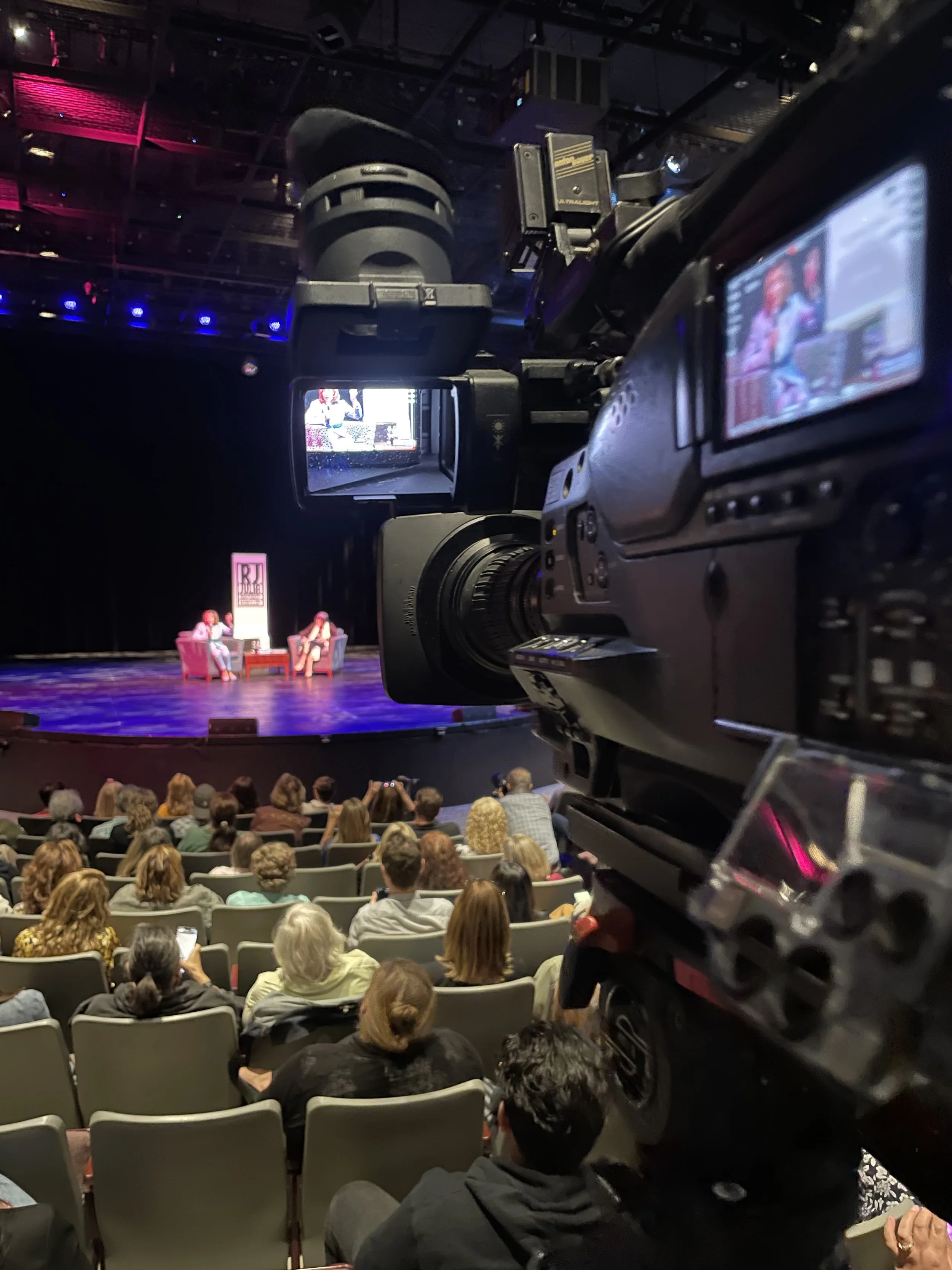 View of an indoor theater or auditorium stage with two women seated on pink armchairs, talking. Audience is seated facing the stage. A large camera is positioned in the foreground capturing the scene.