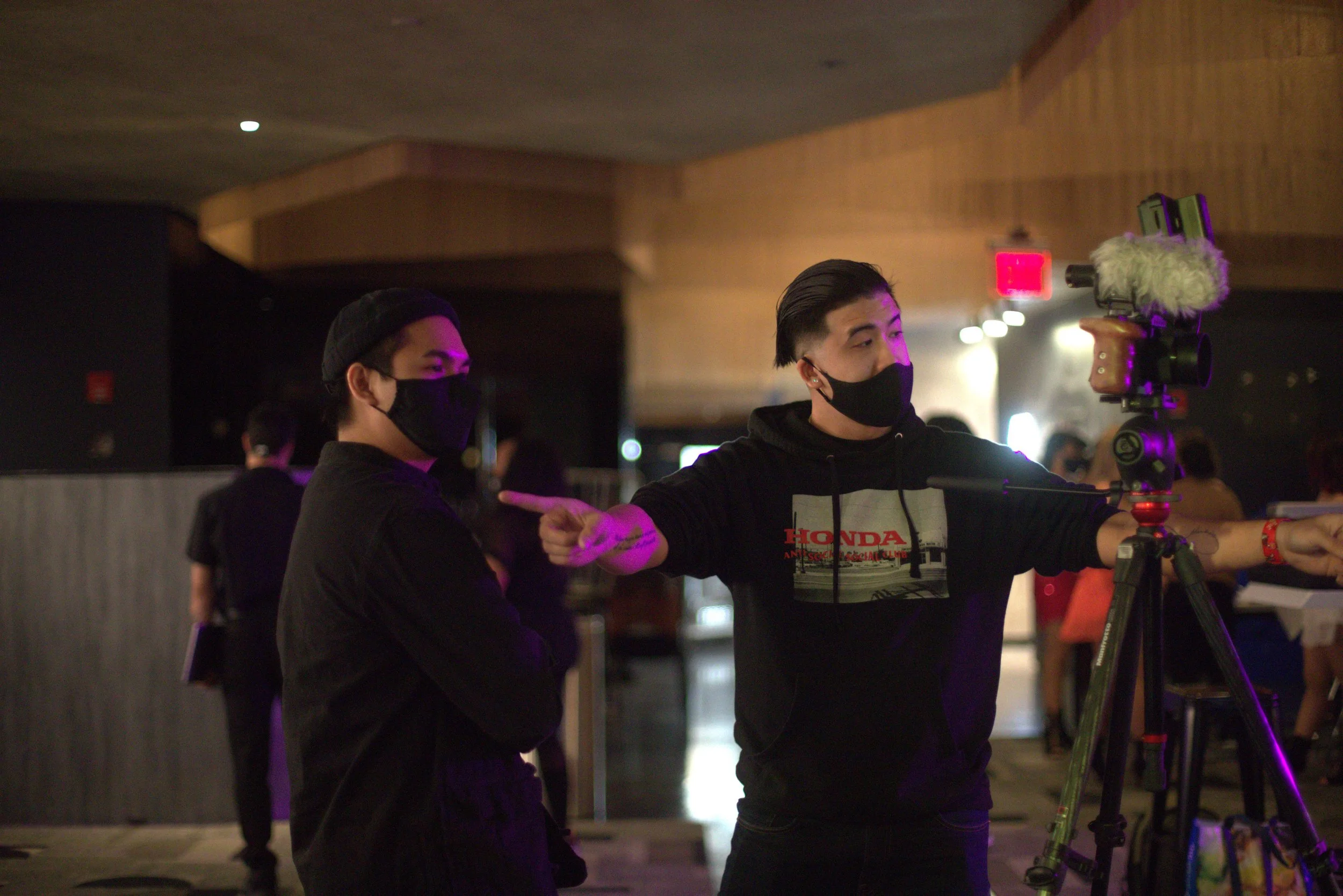 Two young men wearing black face masks are setting up a professional camera on a tripod at an indoor event, with a third person partially visible in the background.