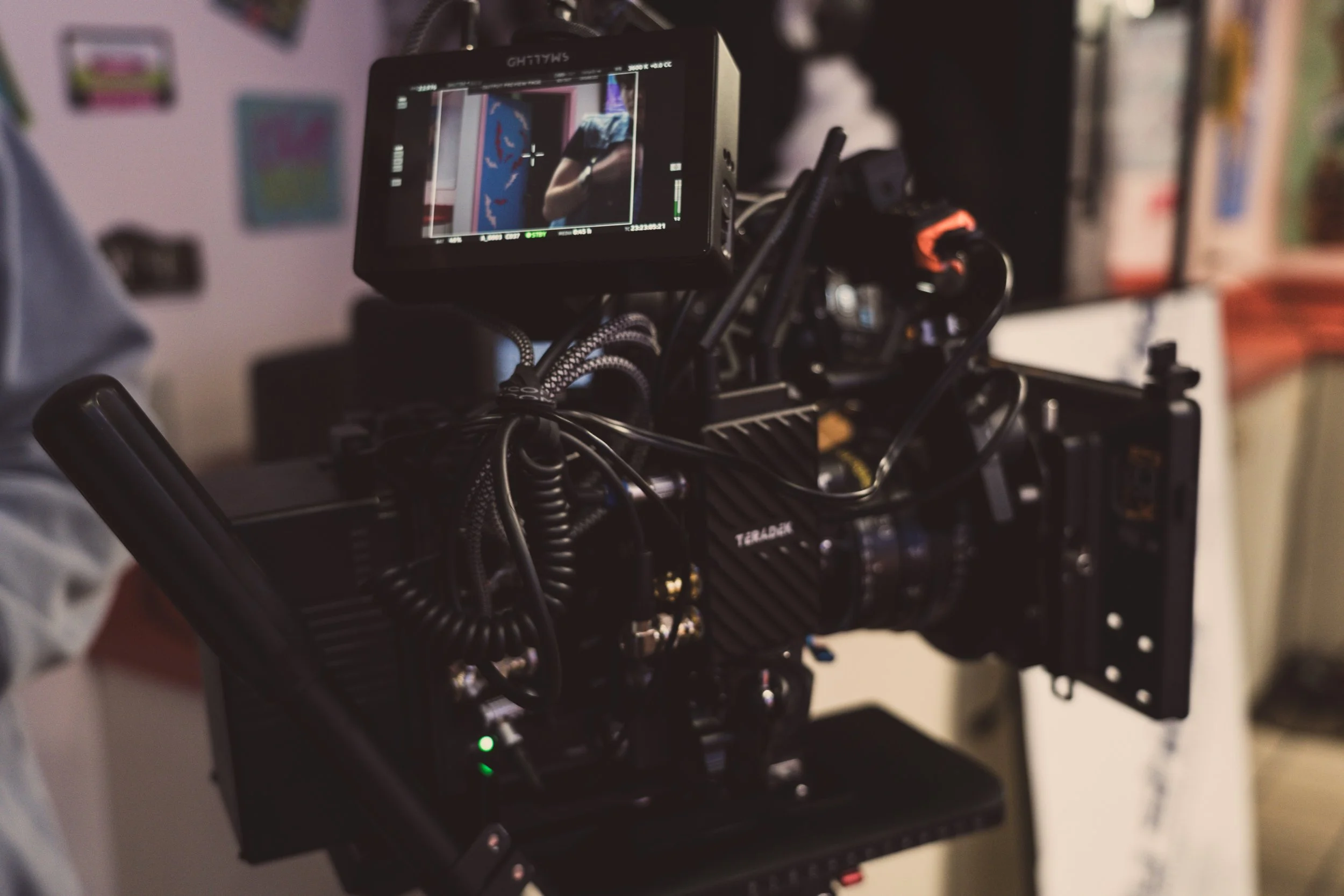 The image shows a professional camera mounted on a rig, capturing footage of a person in a room with colorful wall decorations. The camera's LCD screen reflects the person and the background.