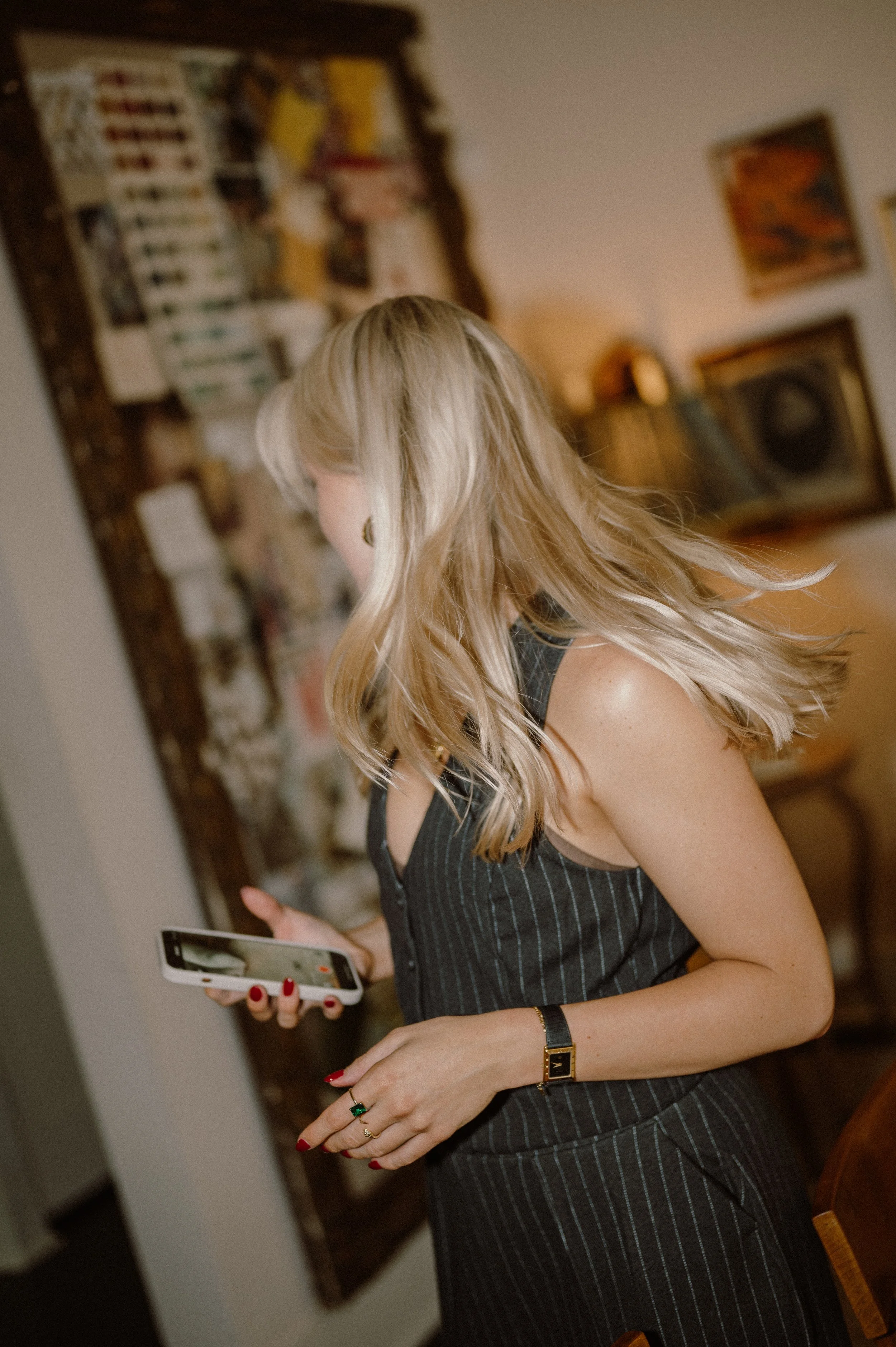 A woman with long blonde hair wearing a black and gray pinstripe dress, holding a smartphone in her left hand, with a jewelry bracelet on her left wrist and rings on her fingers, standing in a room with artwork on the walls and a bookshelf in the background.