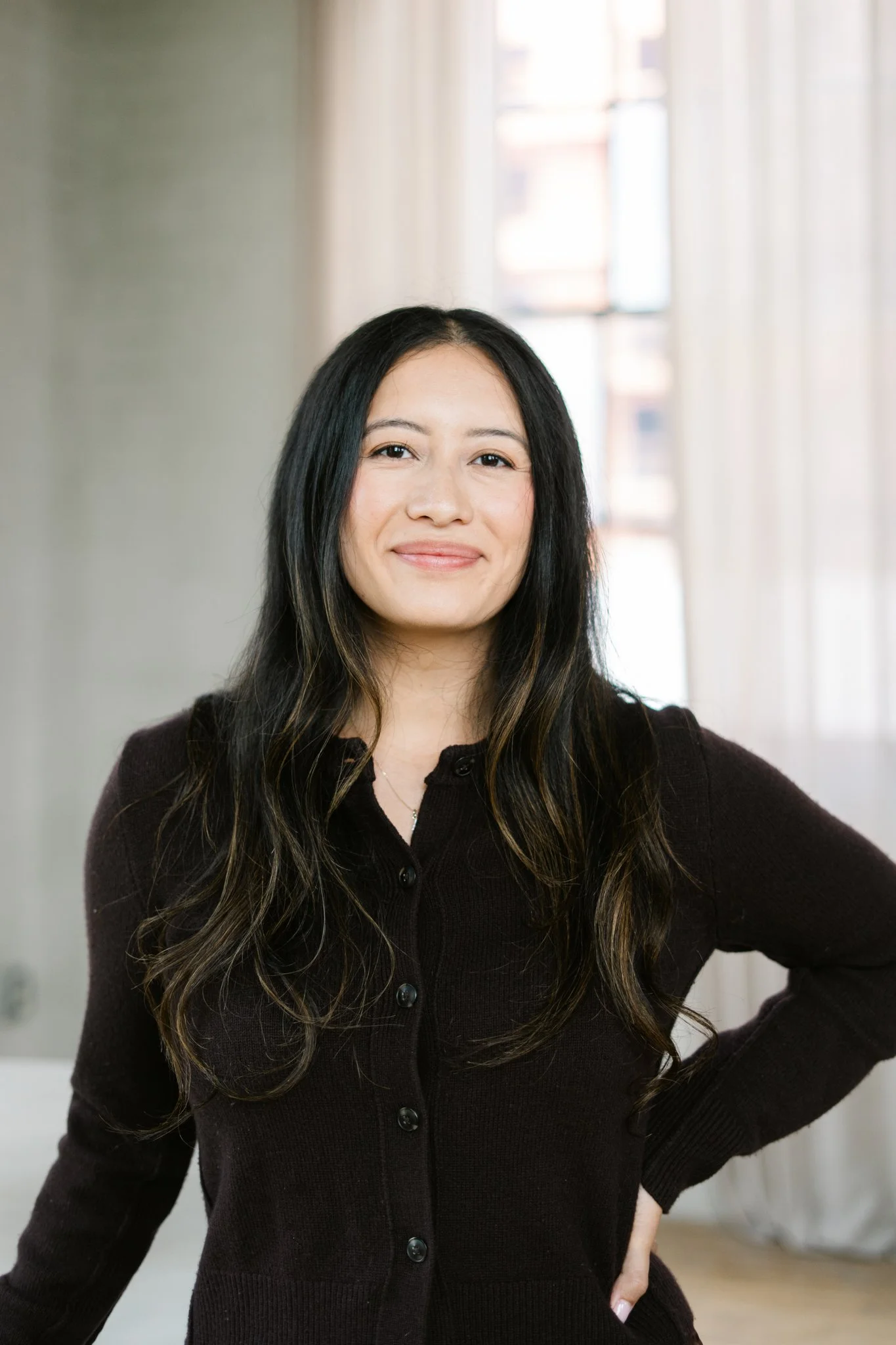 A young woman with long dark hair, wearing a black sleeveless top and light blue ripped jeans, sitting on a beige cushioned bench by a window with white framing in a bright room.