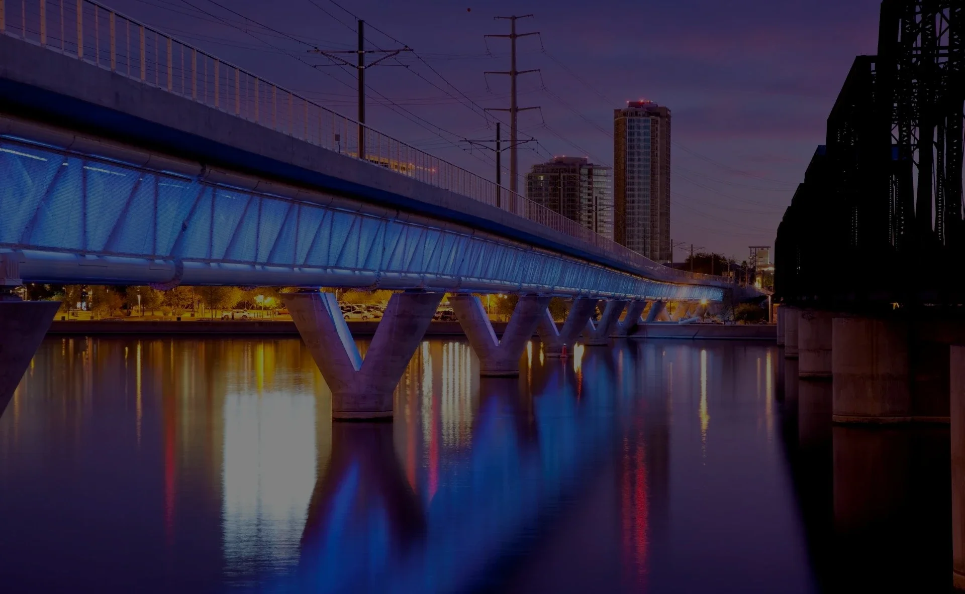 Tempe pedestrian bridge lit with blue lights reflecting over the water at twilight, with city buildings in the background.webp