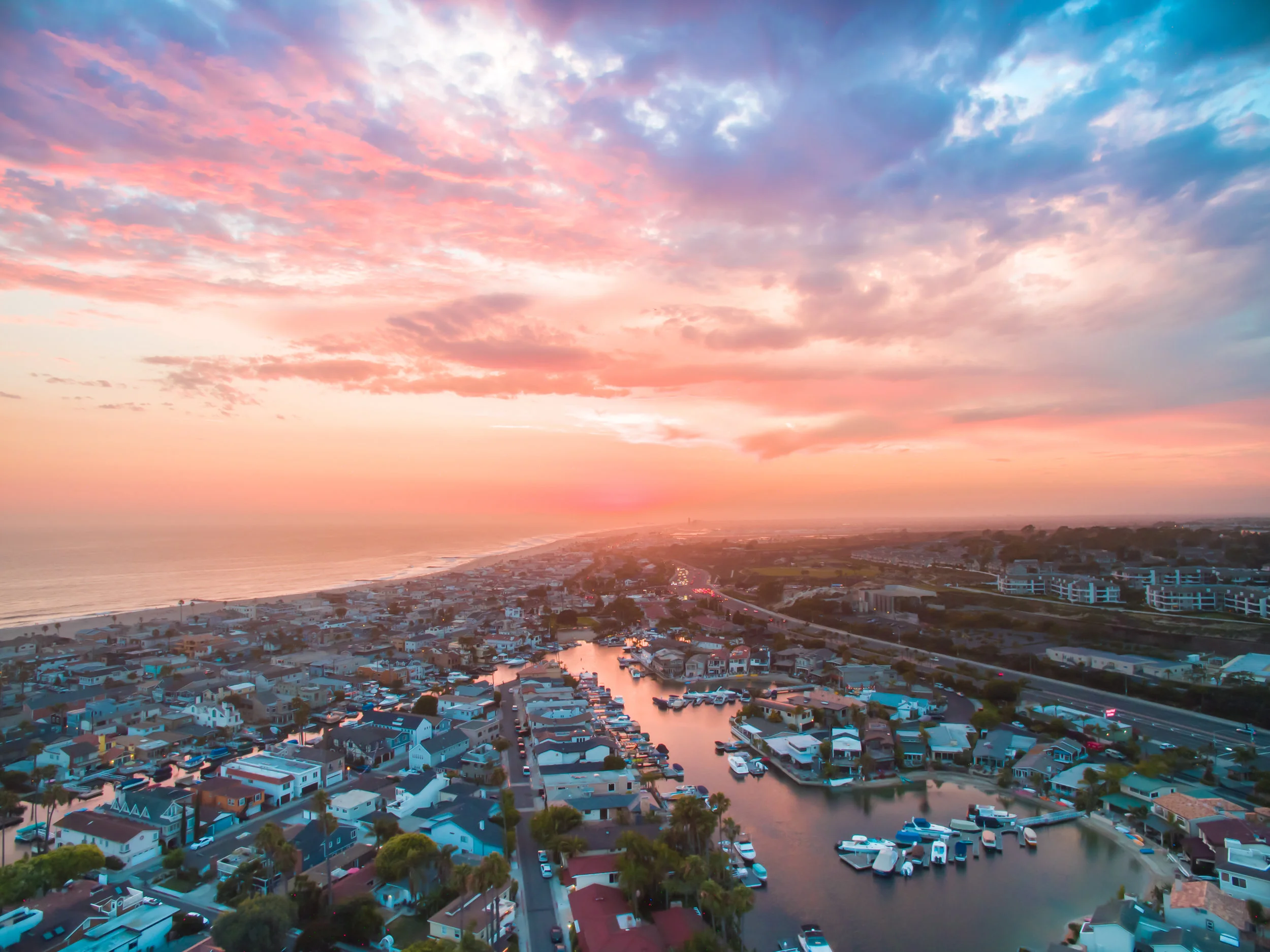 Aerial view of a coastal city at sunset with a harbor filled with boats, residential houses, and a sky with pink and purple clouds.