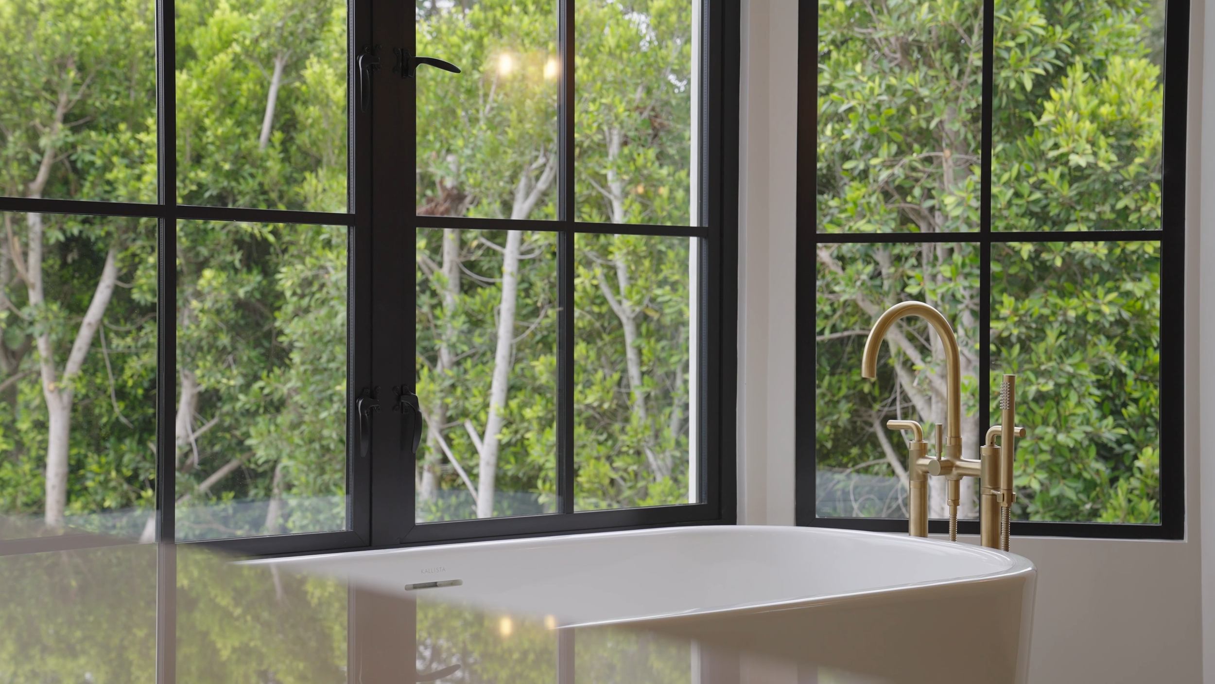 A bathroom with a freestanding bathtub and gold faucet, large windows showing green trees outside.