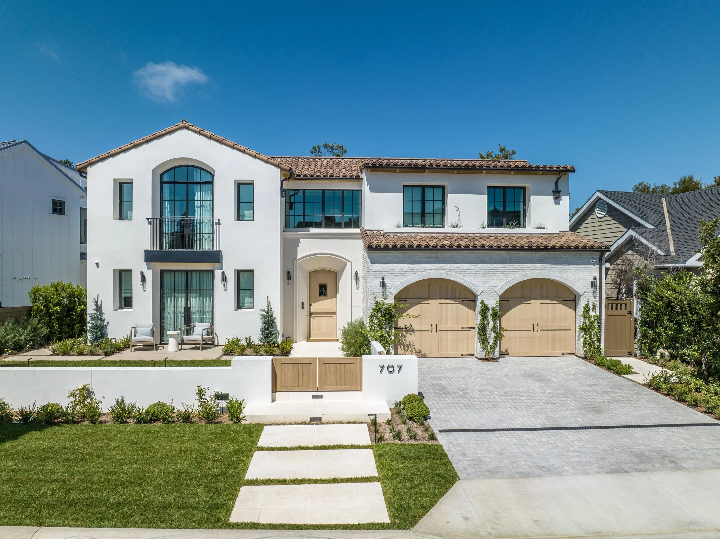 Modern two-story house with a white exterior, terracotta roof tiles, and a landscaped front yard featuring a paved driveway, green lawn, and small bushes. The house has large windows, a small balcony, and a wooden front door.