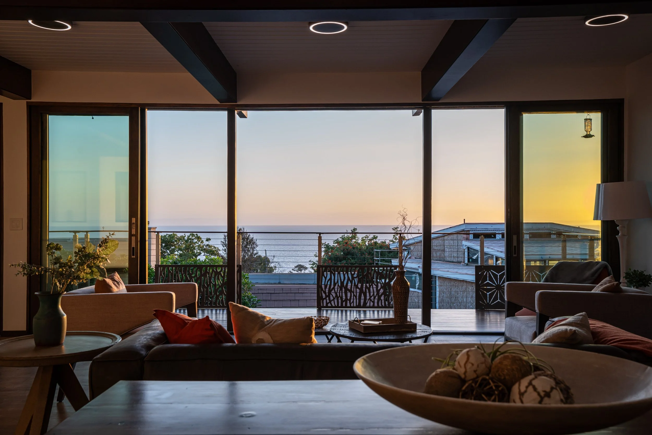 Living room with large glass sliding doors opening to a balcony overlooking the ocean during sunset, with sofas, cushions, and decorative items inside.