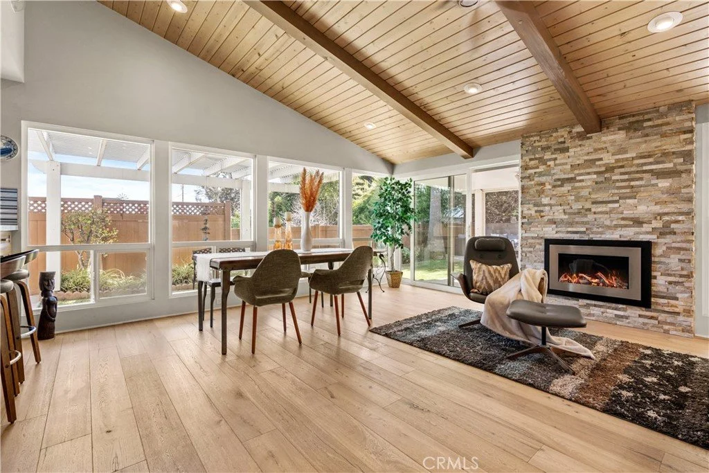 Living room with a wooden ceiling, a stone fireplace, modern chair, dining table with four chairs, large windows, and sliding glass door leading outside.