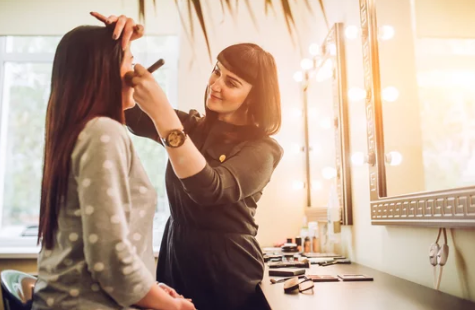 Makeup artist applying makeup to a woman in a dressing room with bright vanity lights.