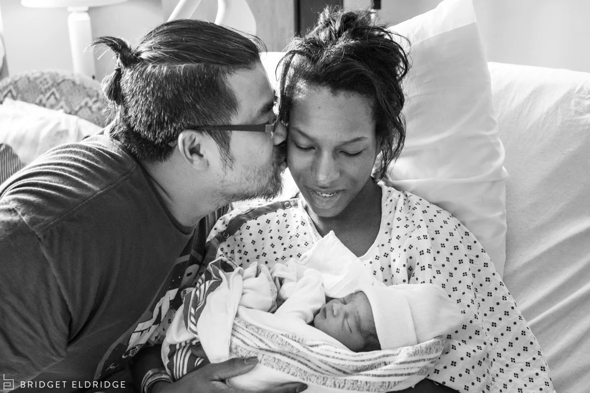 dad kisses mom as she holds her son in the hospital