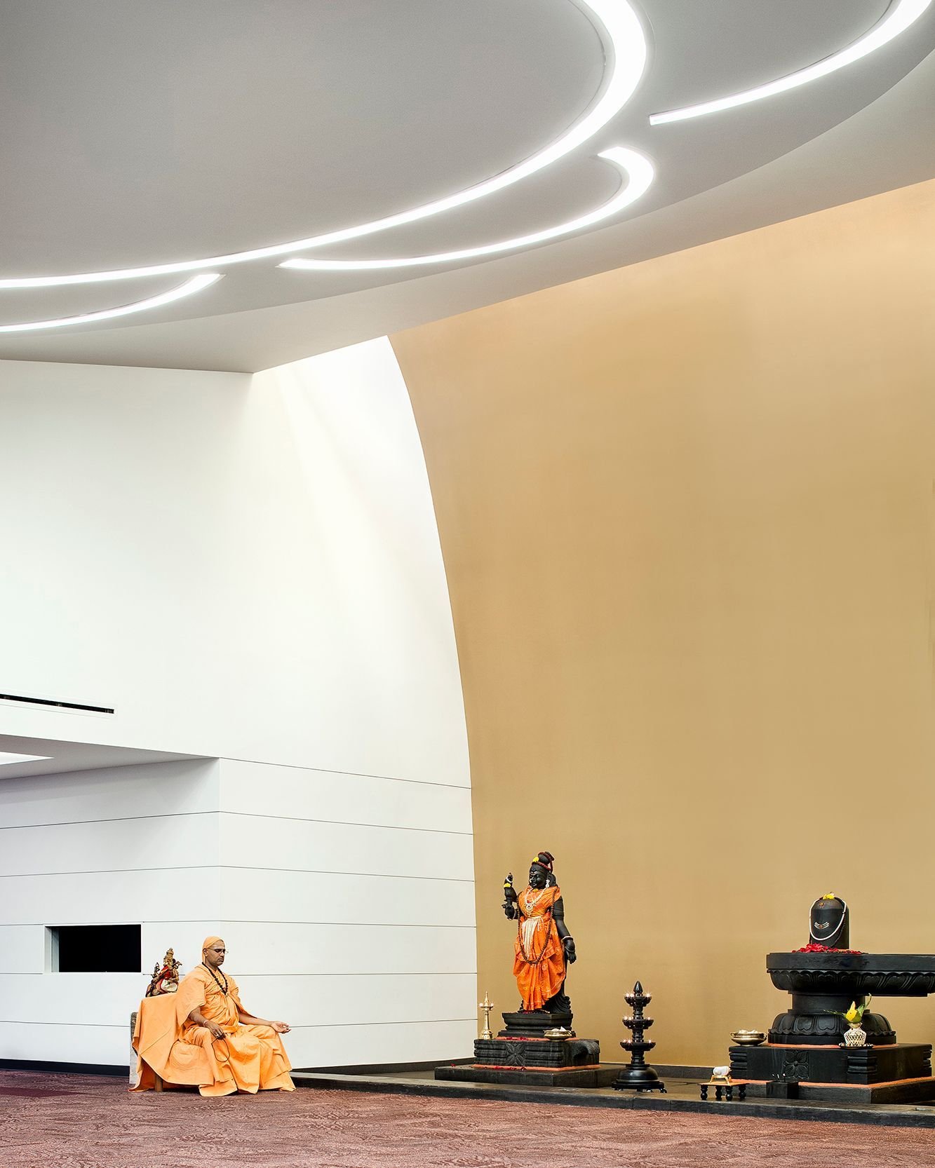 Above the shrine, the most sacred space in the temple, light from concealed skylights is reflected by a golden wall, creating an aura around three deities arrayed with bright robes and flowers. #ChinmayaMissionAustin #MiroRiveraArchitects #MRAProject