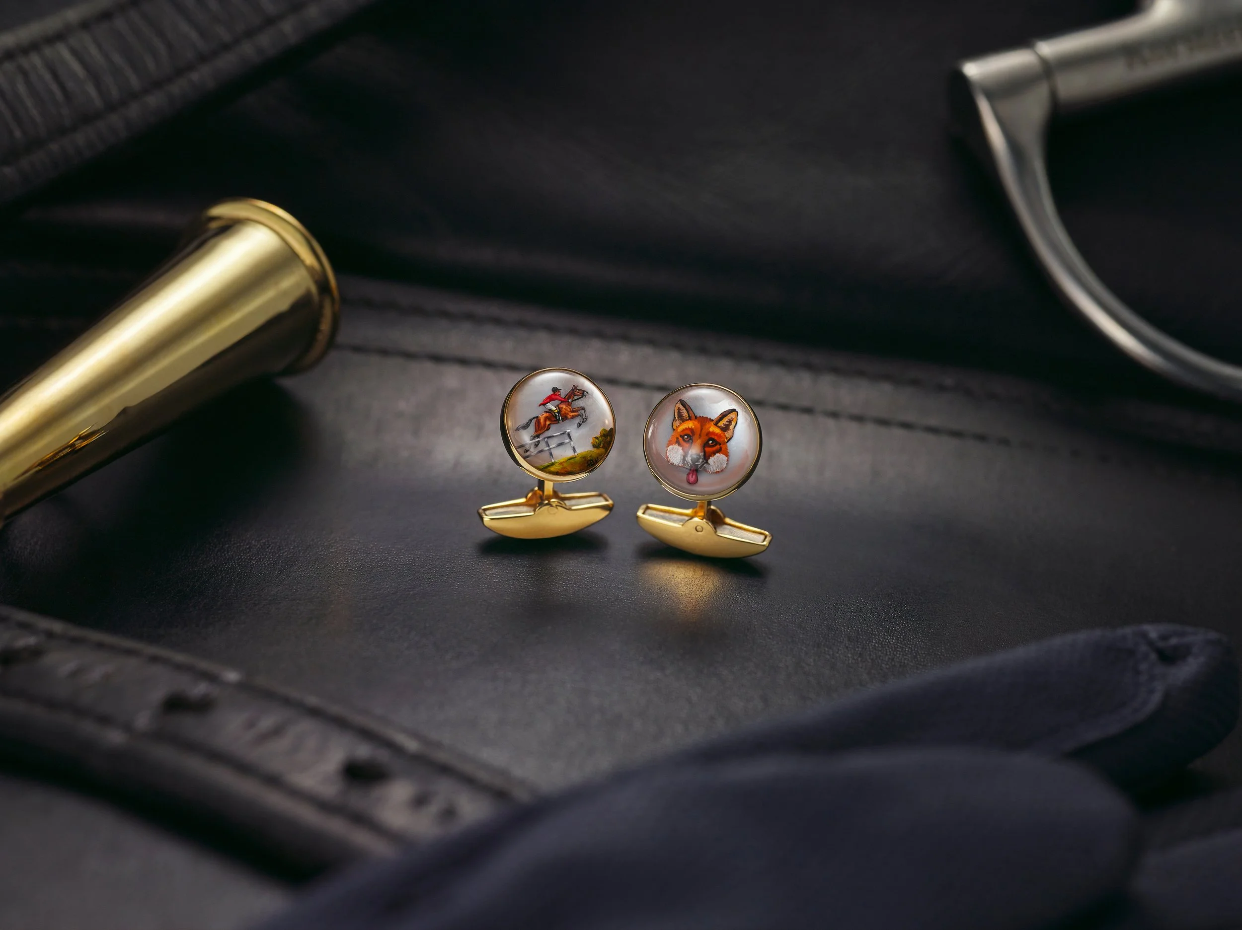 Close-up of decorative cufflinks featuring a jockey on a horse and a fox's face, placed in a black case with a black background and a metallic pen nearby.