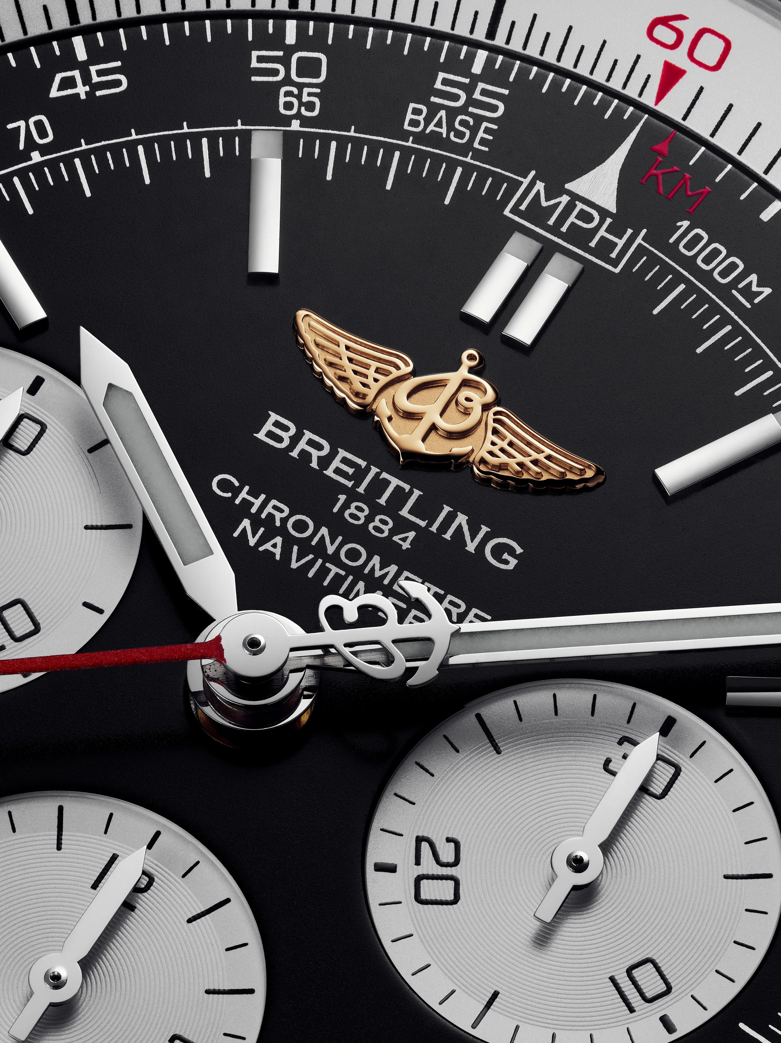 Close-up of a Breitling chronograph watch face showing multiple subdials, white hour and minute hands, a red second hand, and branding. The watch features a black background with white and gold accents.