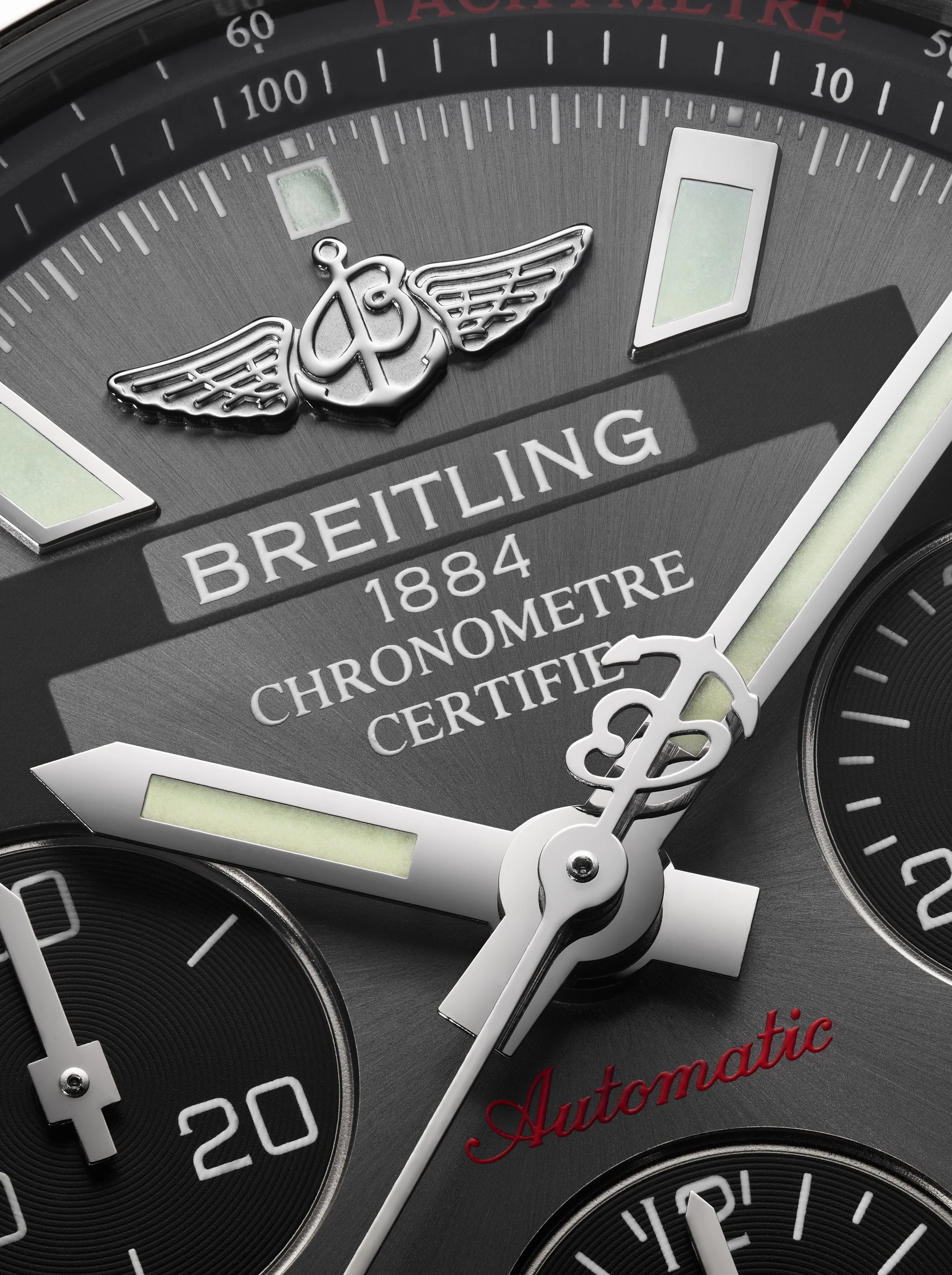 A close-up of a wristwatch face featuring a winged logo, the word 'Breitling,' and details about chronometer certification. The watch has a dark background with white and metallic accents, luminous markers, and multiple subdials.