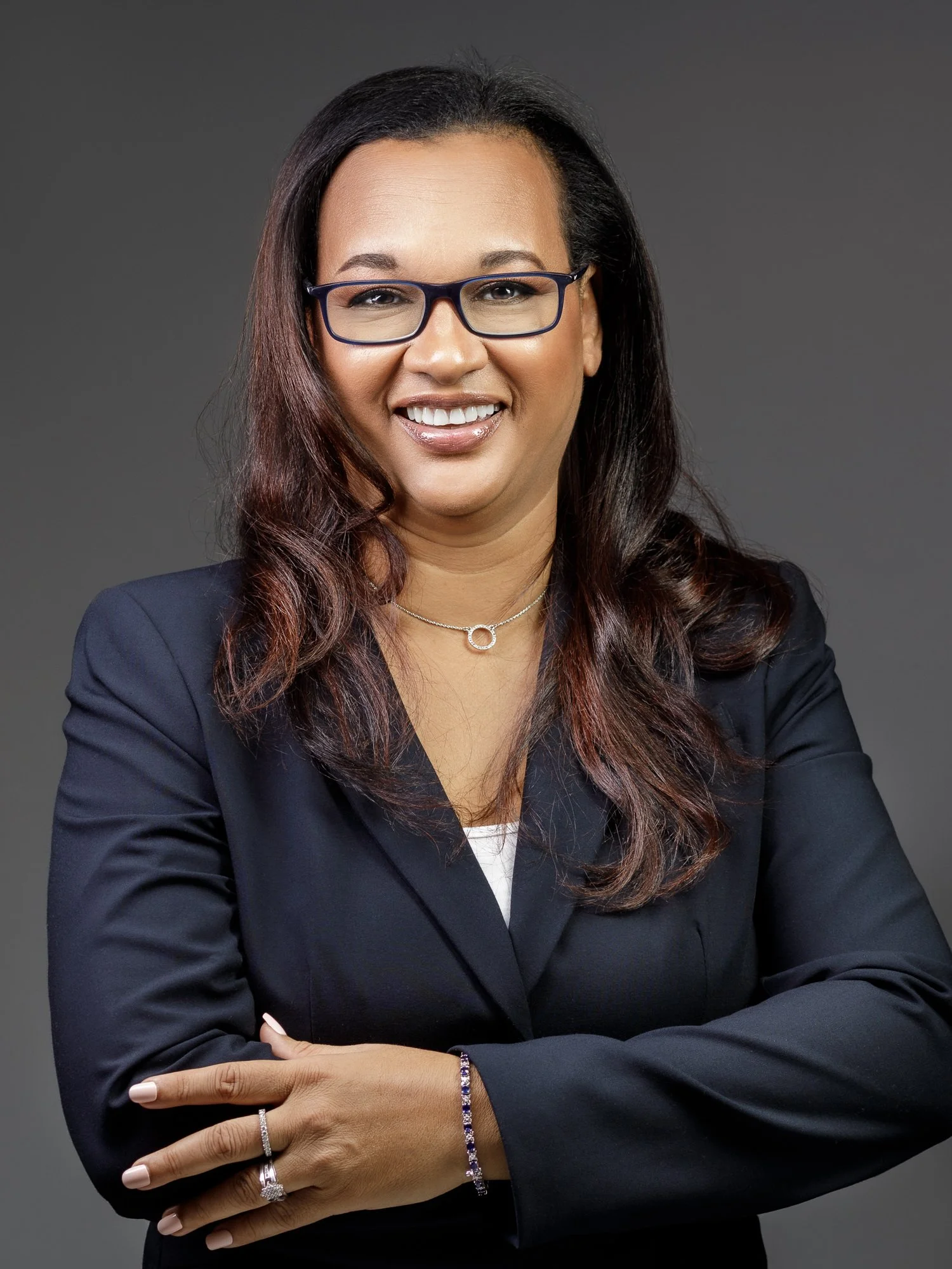 A confident businesswoman in a dark blazer and glasses, smiling with arms crossed against a gray background.